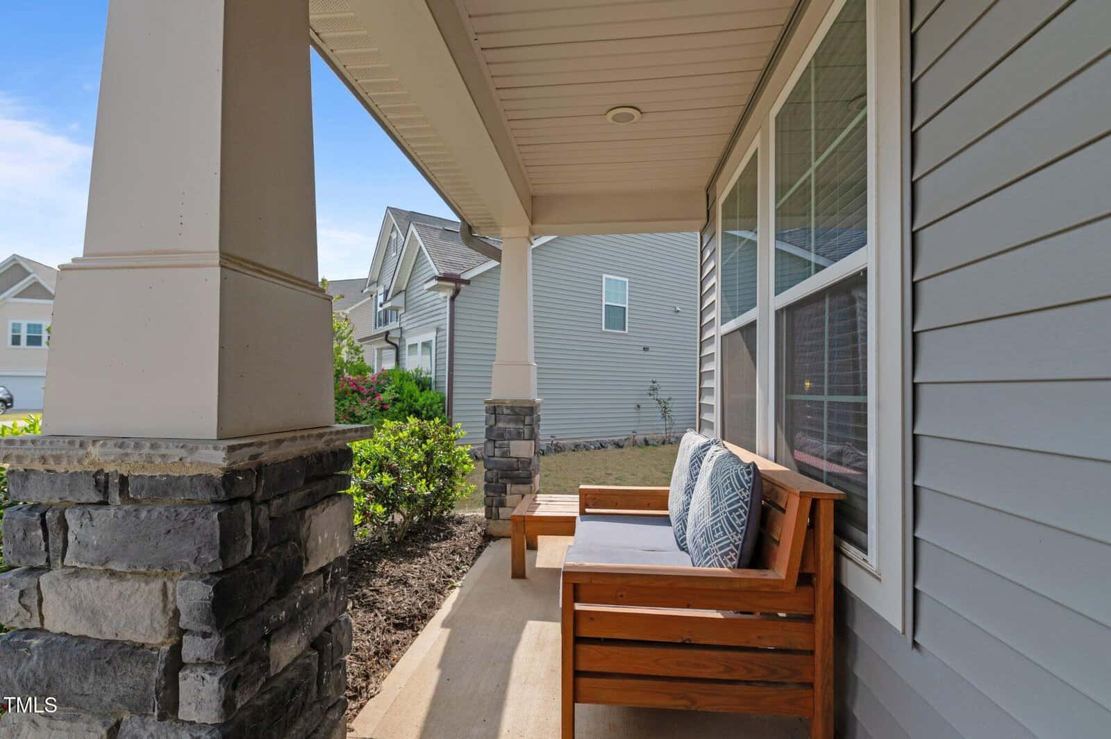 A cozy wooden bench with cushions sits on a covered front porch of a modern house, featuring stone columns and gray siding. The porch overlooks a small garden and neighboring homes on a sunny day.