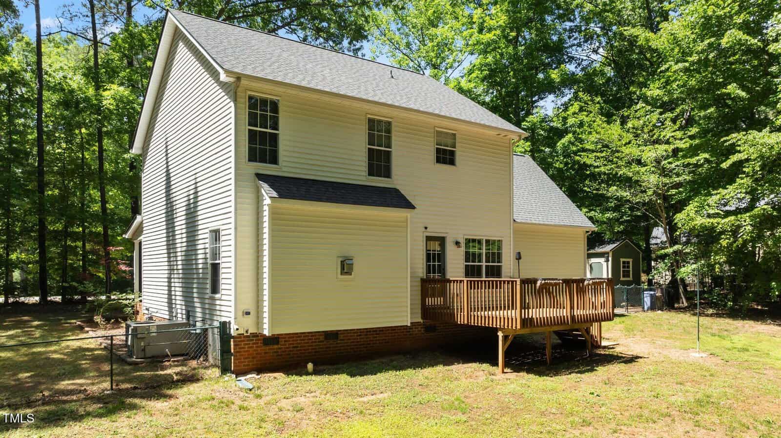 Two-story white house with gray roof, multiple windows, and a wooden deck in the backyard. The yard is partly shaded by tall trees and has patches of grass and bare ground.