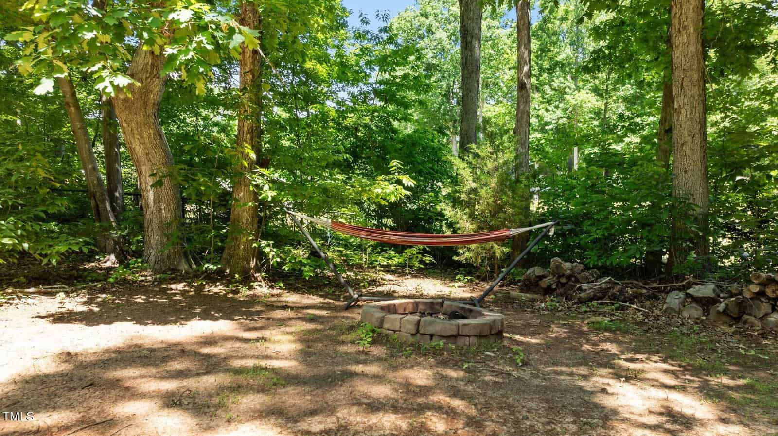 A striped hammock is suspended above a circular stone fire pit in a shaded, wooded backyard with scattered logs and dense green foliage.