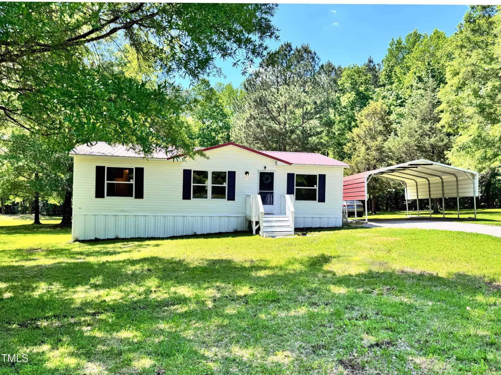 A single-story white manufactured home with black shutters, a red roof, and front steps sits on a grassy lawn at 6579 Sidney Cottrell Road, Oxford, with trees behind and a metal carport to the right.