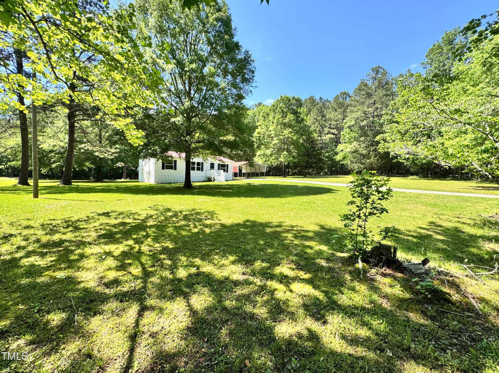 A white single-story house with a red roof sits on a large grassy lawn surrounded by trees under a clear blue sky. Sunlight creates shadows from the trees across the yard.