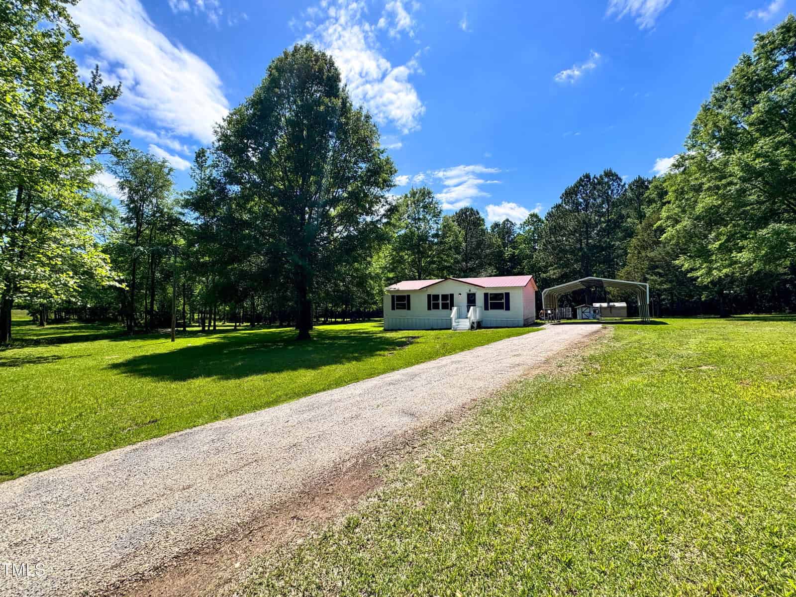 A small white house with a red roof sits at the end of a gravel driveway, surrounded by green grass and large trees, with a metal carport on the right under a blue sky with scattered clouds.