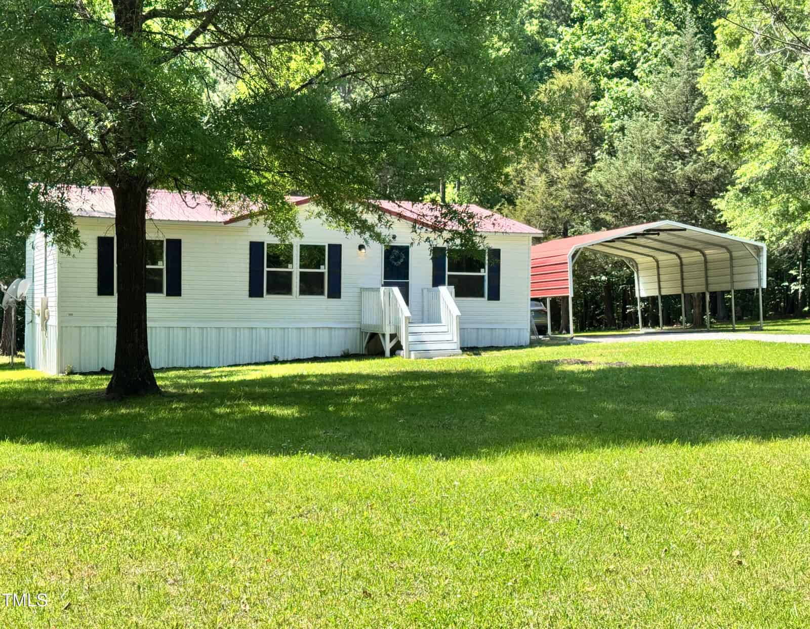 A white manufactured home with black shutters and a red roof sits on a grassy lawn next to a metal carport, surrounded by trees under bright daylight.
