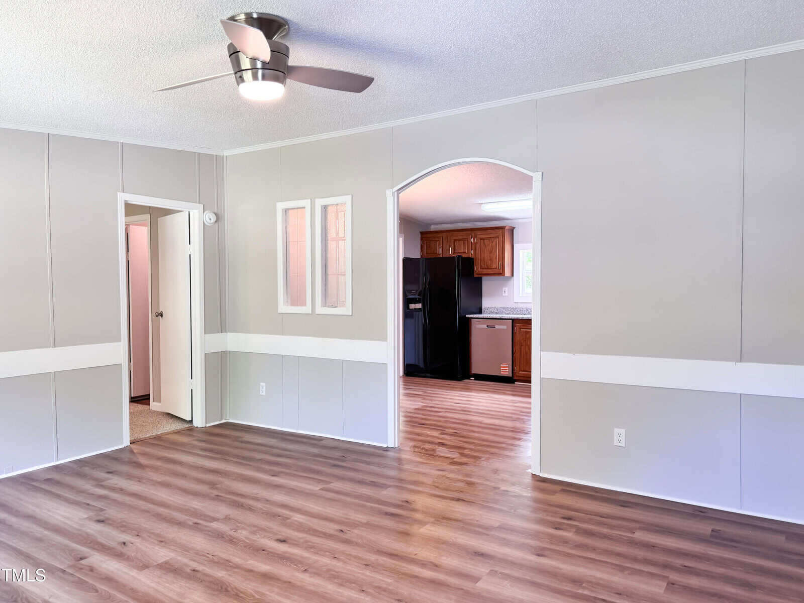 A room with light gray walls, wood-look flooring, a ceiling fan, and white trim. An open doorway leads to a kitchen with wood cabinets and stainless steel appliances. Another doorway leads to a carpeted room.