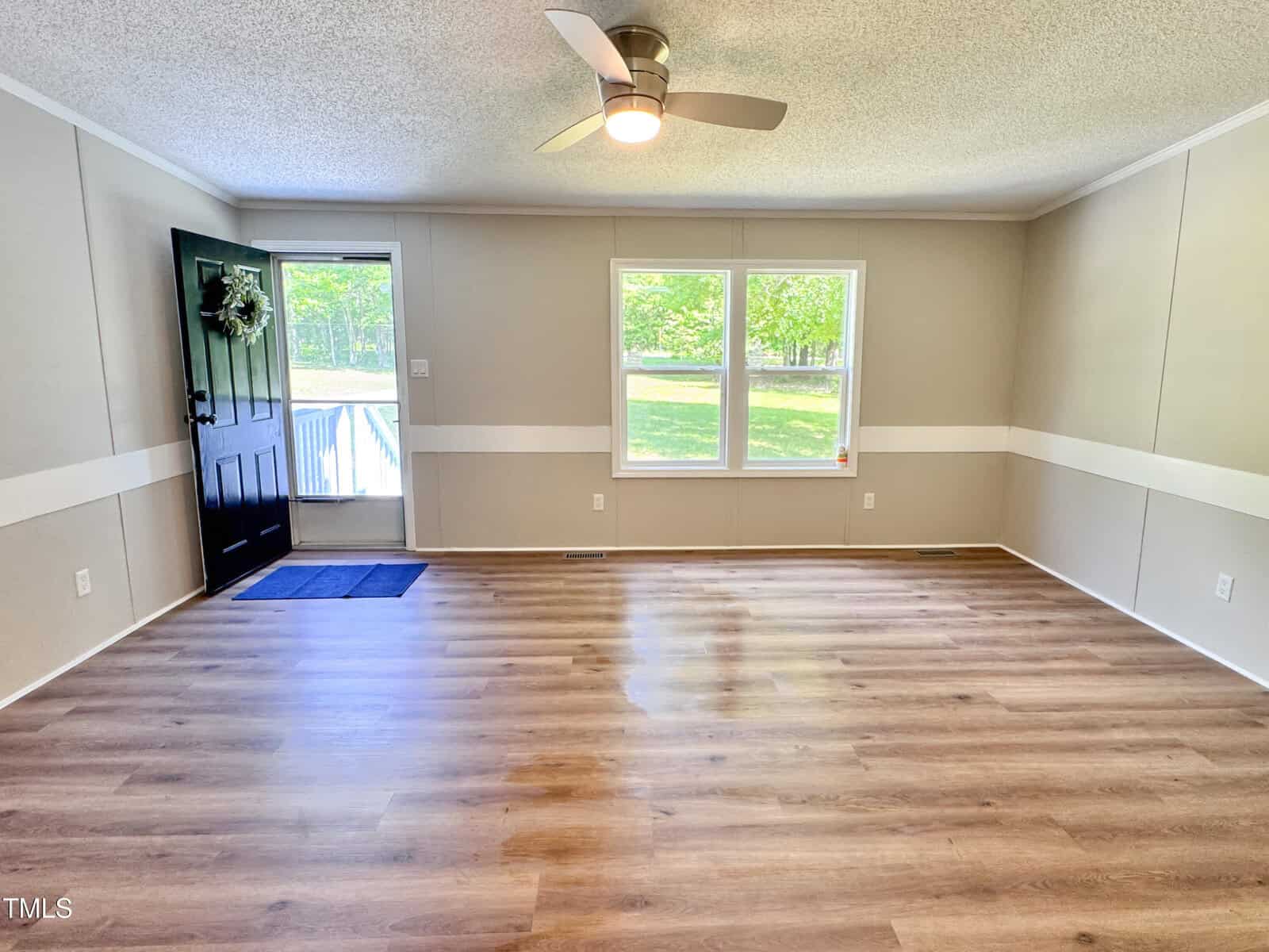 Unfurnished living room with light wood flooring, beige walls with a white stripe, a ceiling fan with light, a front door with a wreath, large window, and natural light coming in.