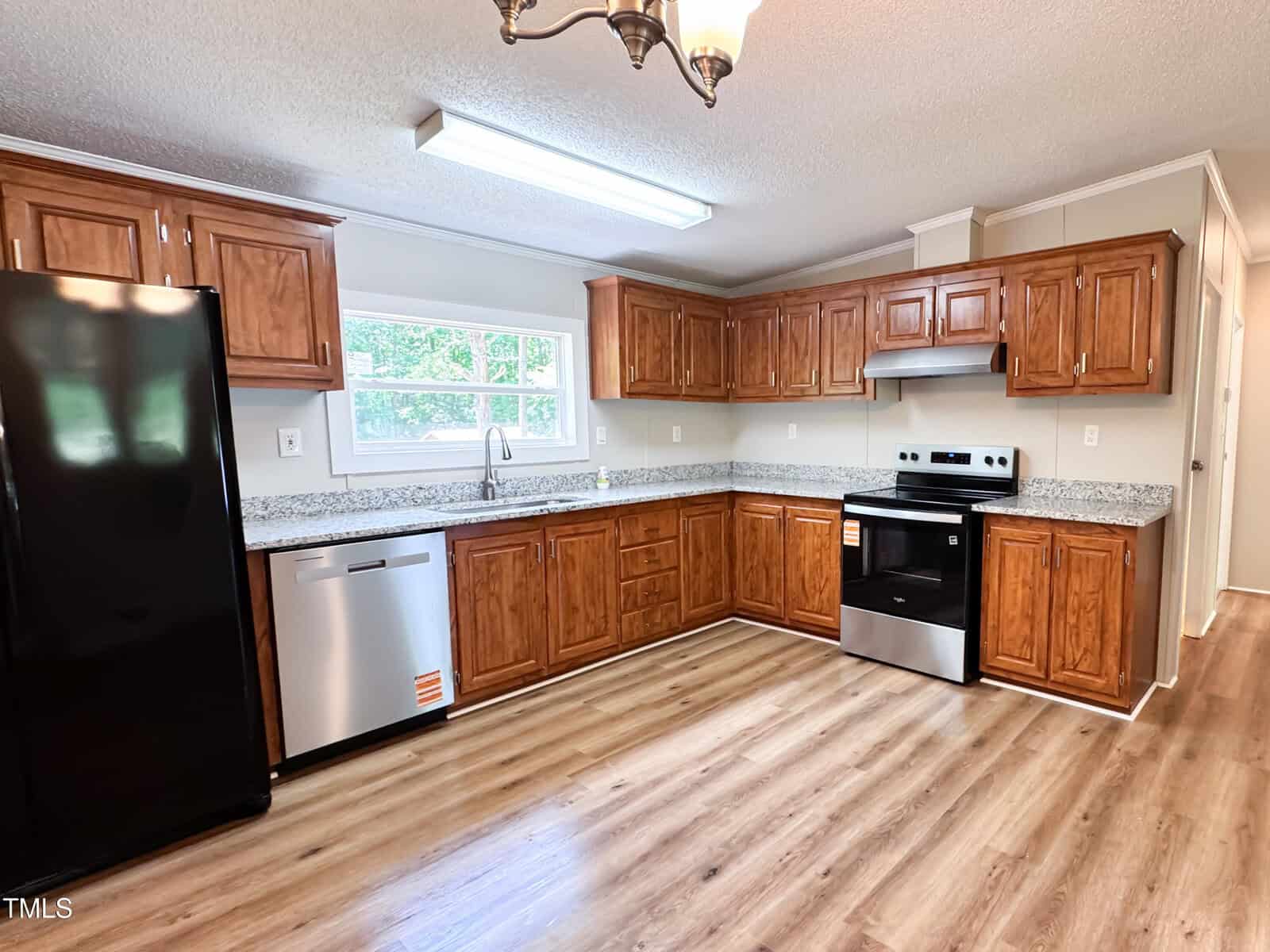 A kitchen with wooden cabinets, granite countertops, stainless steel appliances including a stove and dishwasher, a black refrigerator, a window above the sink, and wood-patterned flooring.