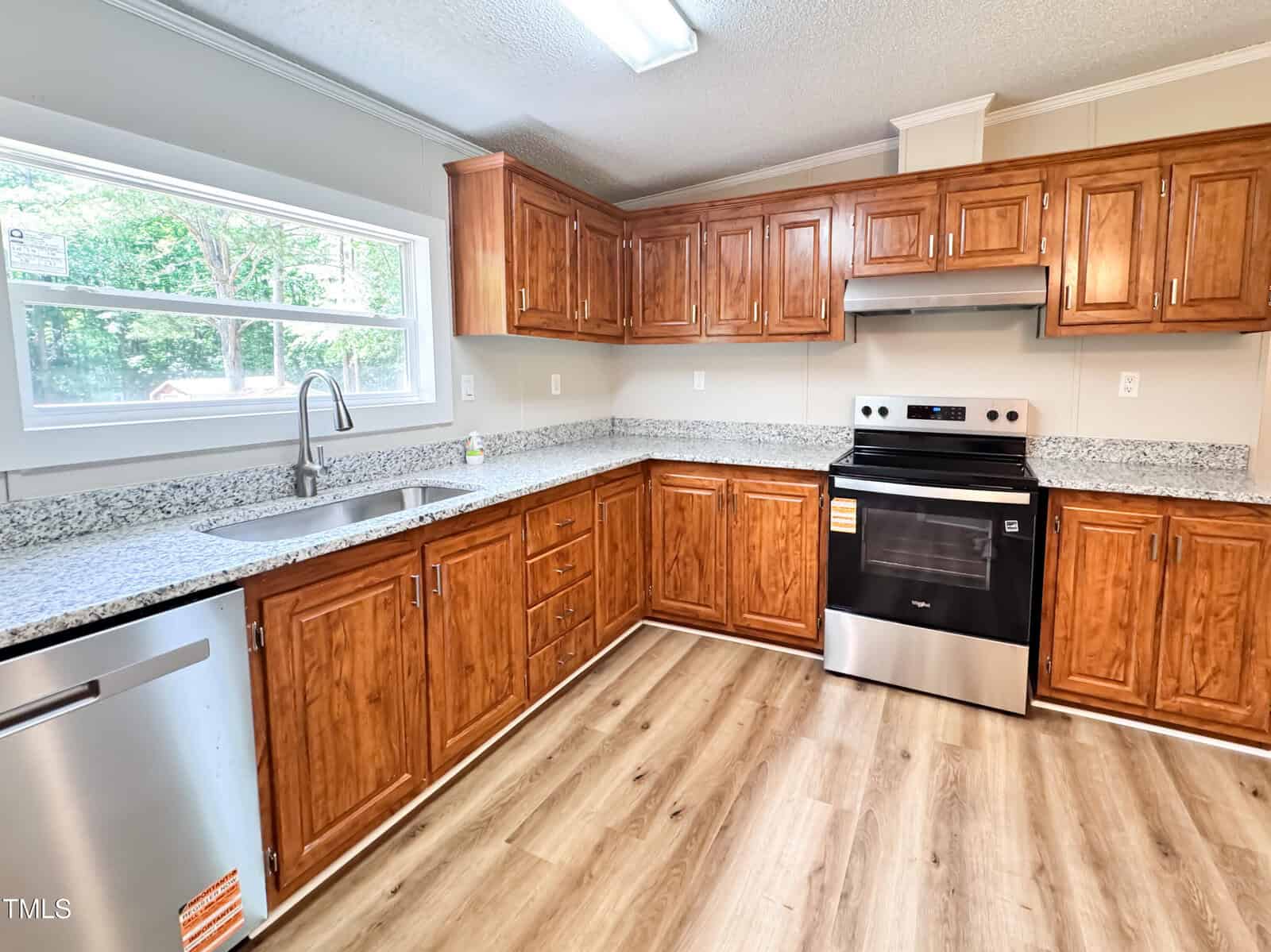 A modern kitchen with wood cabinets, granite countertops, stainless steel appliances including a stove and dishwasher, a large window above the sink, and light wood flooring.
