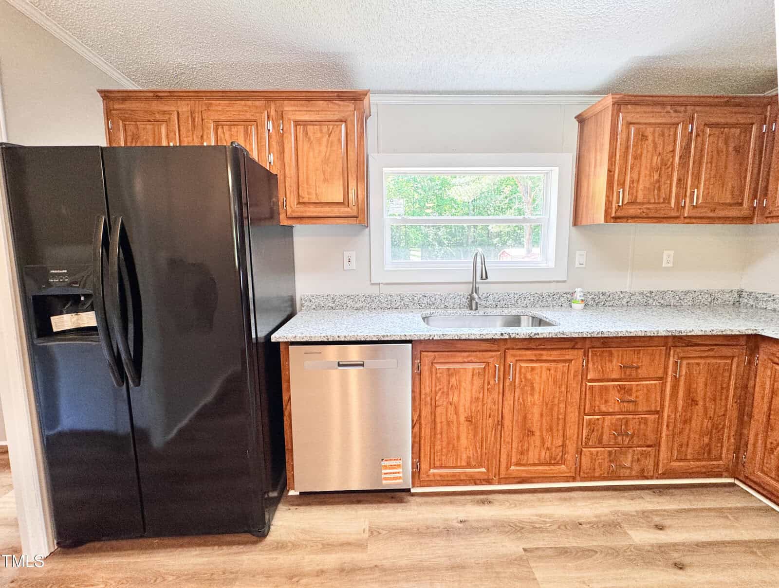 A kitchen with wooden cabinets, a black refrigerator, a stainless steel dishwasher, a stainless steel sink under a window, granite countertops, and light wood flooring.
