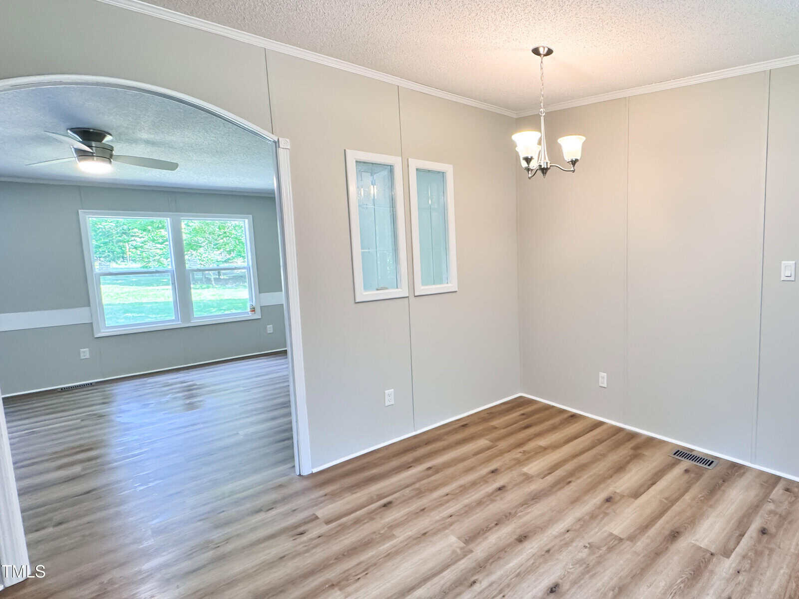A dining area with light wood flooring, beige walls, a chandelier, two glass display cabinets, and an open doorway leading to a living room with large windows, ceiling fan, and similar flooring.