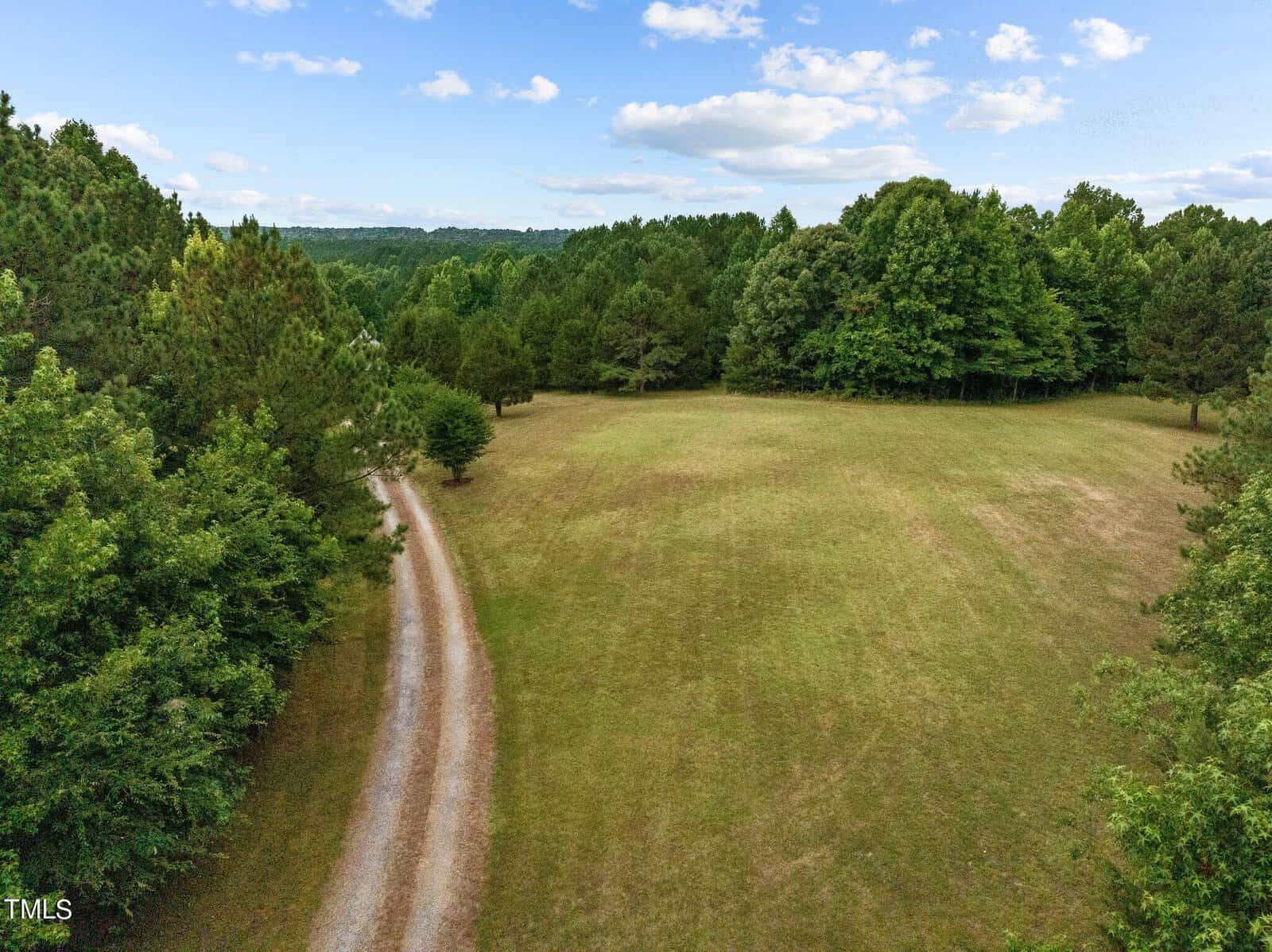 A gravel driveway curves through a grassy clearing surrounded by dense green trees under a blue sky with scattered clouds.