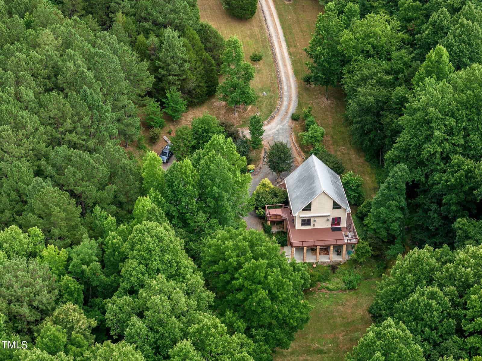 Aerial view of a two-story house with a porch, surrounded by dense green trees and a grassy yard. A gravel driveway curves around the property, leading to a parked vehicle partially visible in the trees.