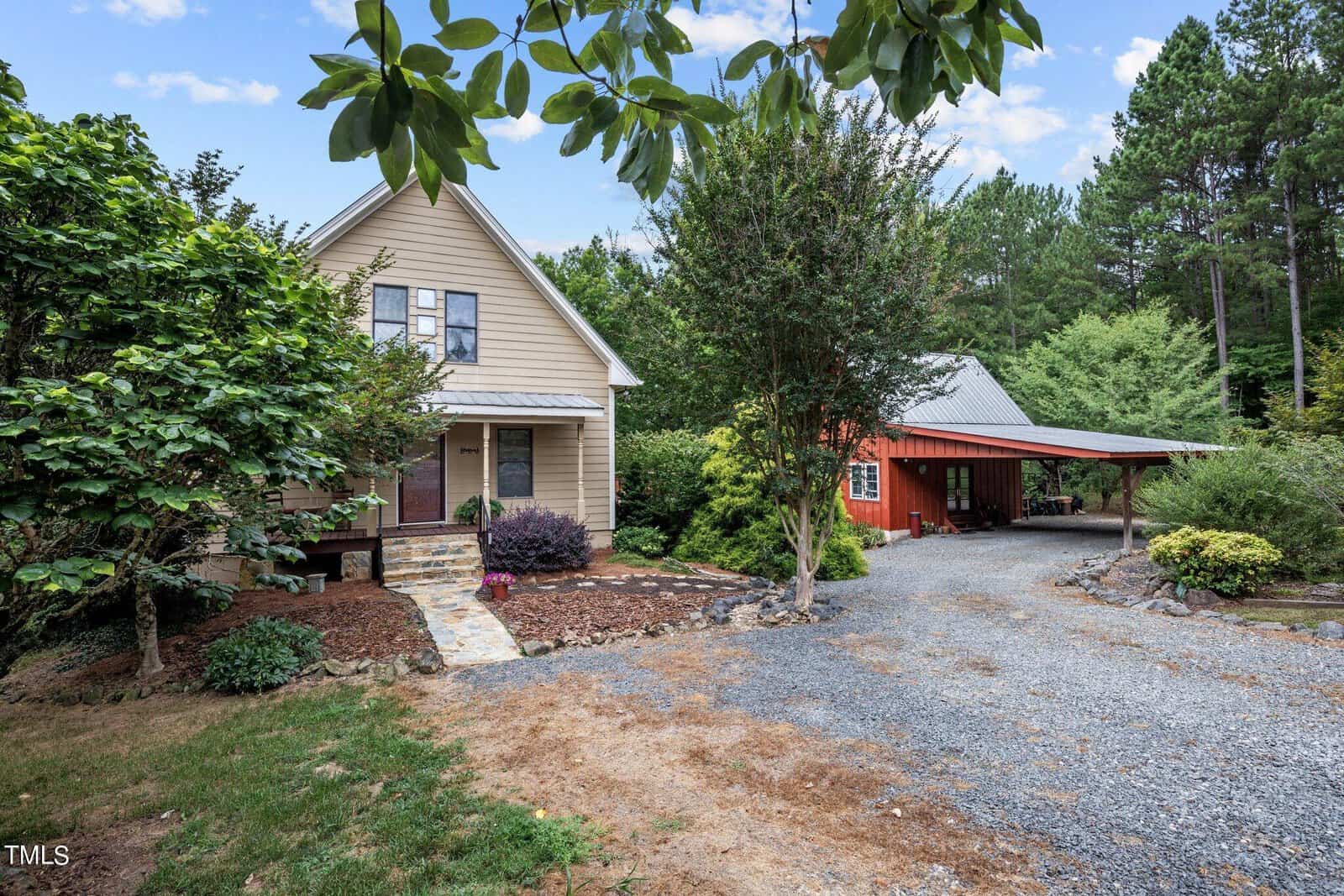 A beige house with a front porch and gable roof sits next to a red barn-style carport surrounded by trees and a gravel driveway under a partly cloudy sky.