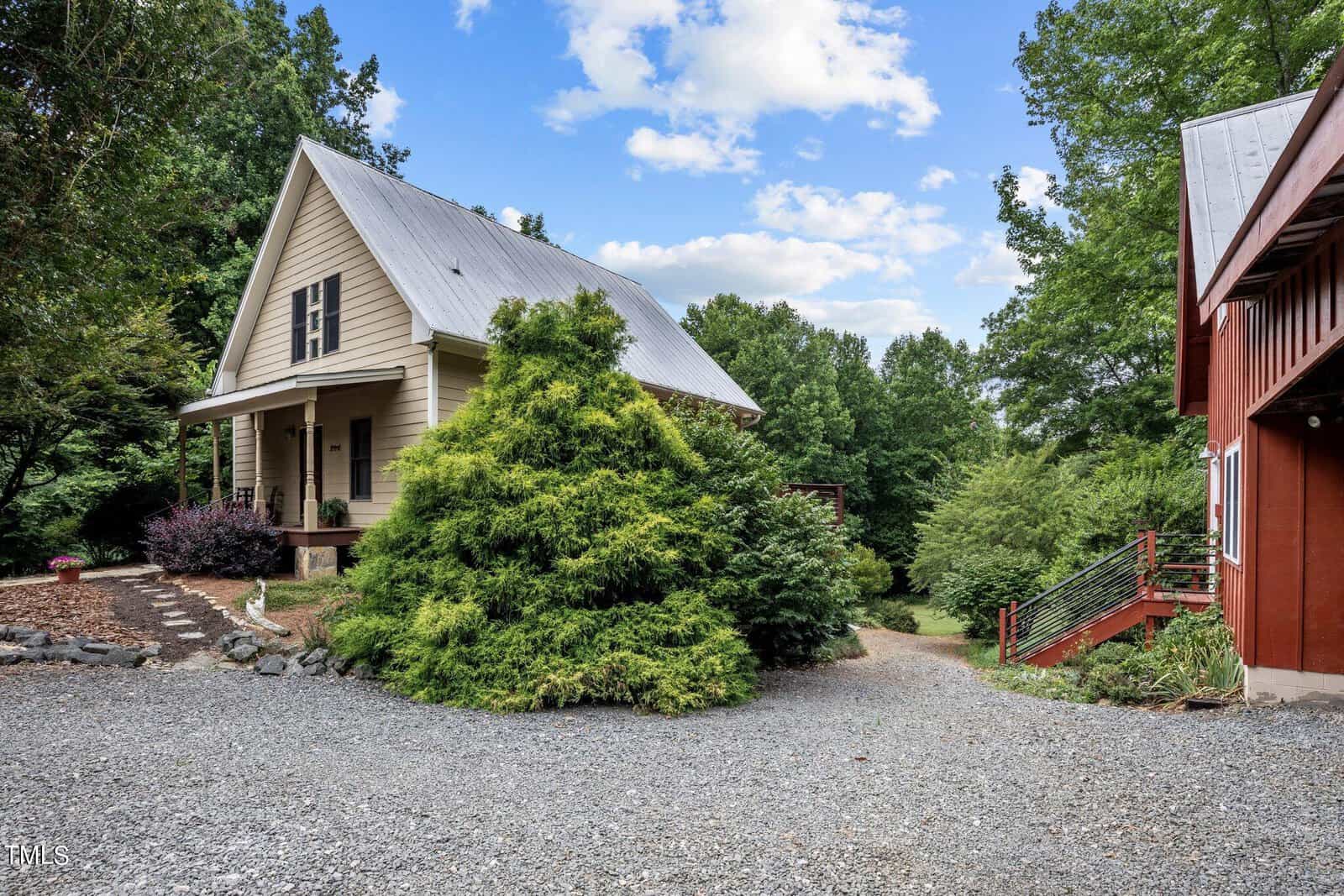 A small beige house with a metal roof stands behind a large green bush. A red building with a staircase is visible on the right. The foreground features a gravel driveway; trees fill the background under a partly cloudy sky.