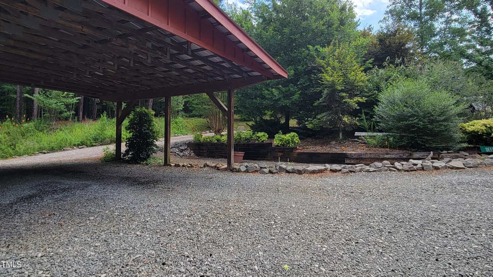 A gravel driveway curves around a covered carport with a wooden roof. There are landscaped garden beds with rocks and various trees and shrubs in the background. The scene is outdoors with natural greenery.