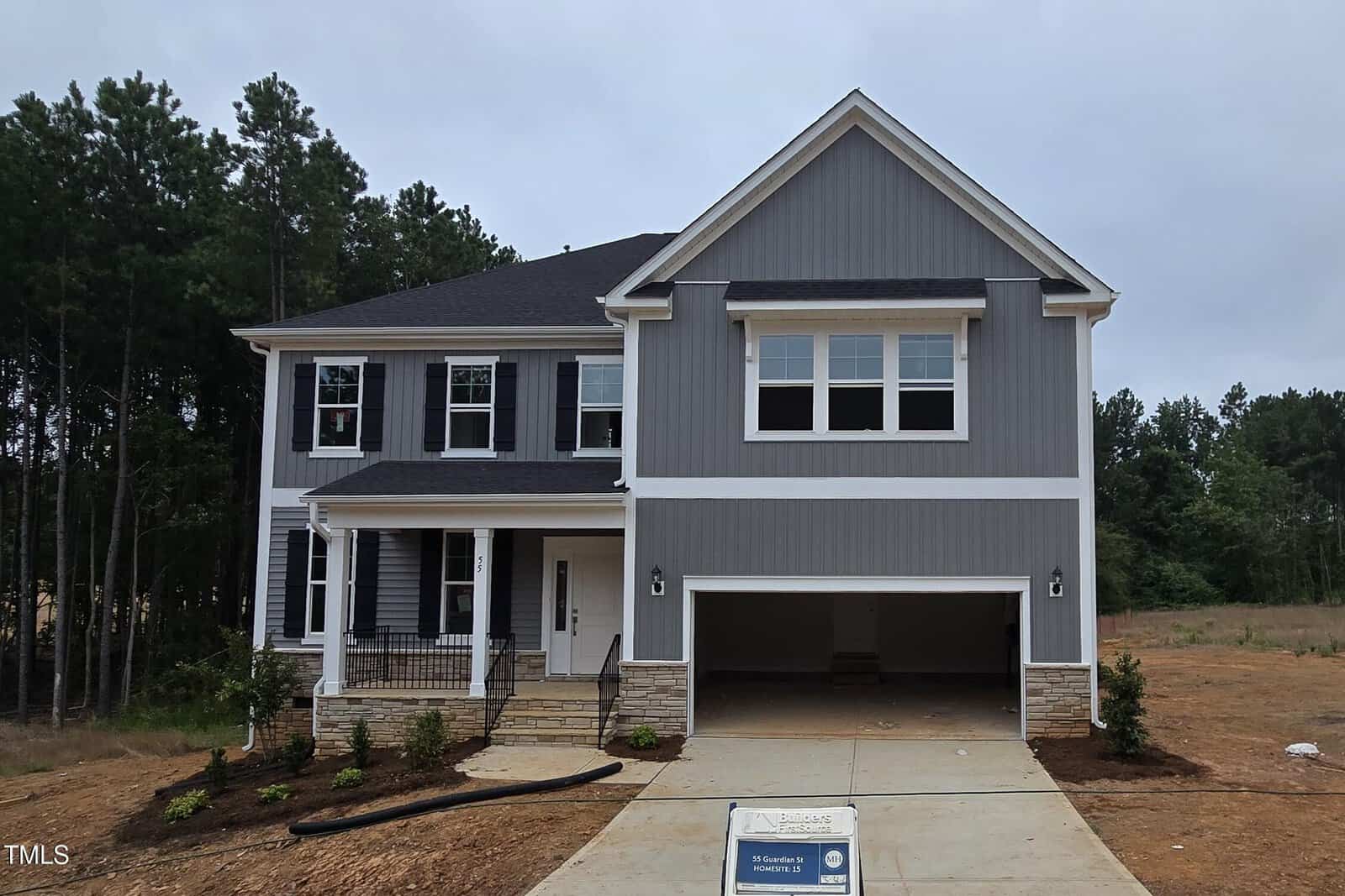 A two-story modern house with gray siding, white trim, and a covered porch. The garage door is open, and the driveway is unfinished. Young shrubs line the front walkway; tall trees are in the background under a cloudy sky.