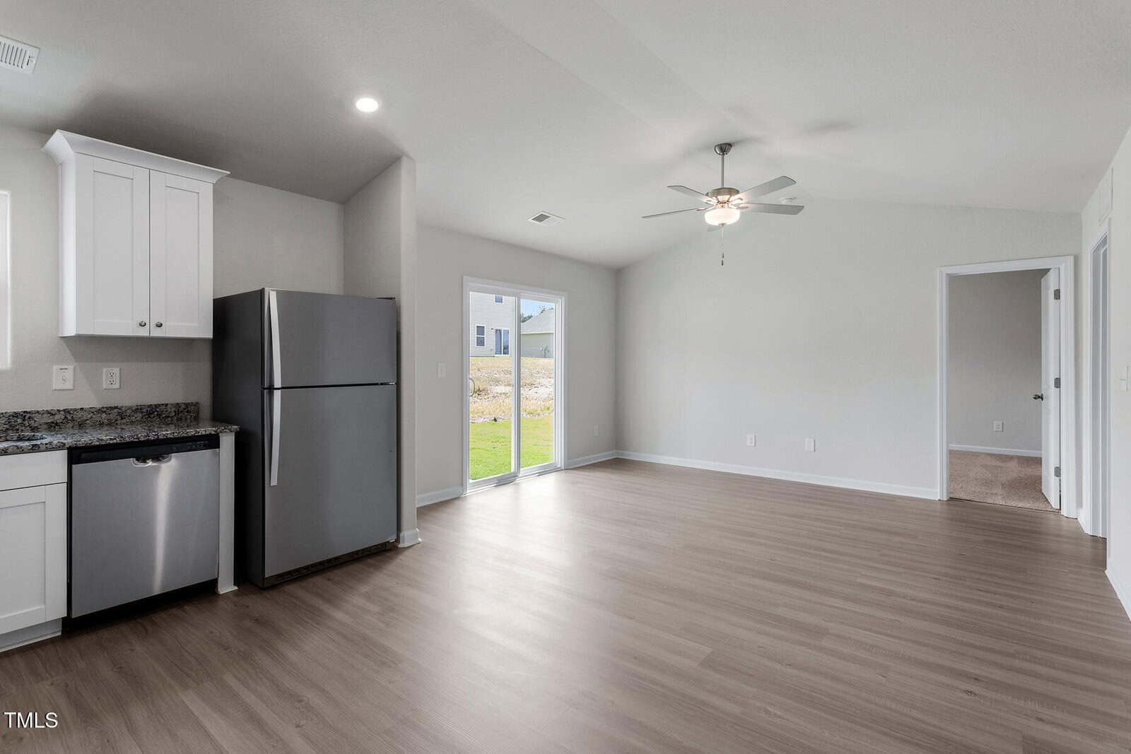 A modern, unfurnished kitchen and living area with light wood floors, white cabinets, stainless steel appliances, a ceiling fan, sliding glass doors, and an adjacent room visible through an open doorway.