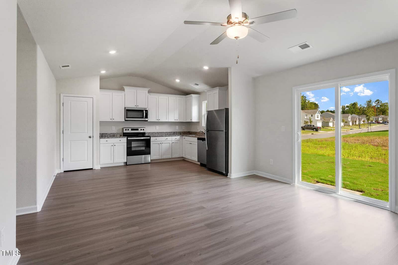 Open-plan kitchen and living area with white cabinets, stainless steel appliances, light wood flooring, ceiling fan, and large sliding glass doors overlooking a grassy suburban street with houses and blue sky.