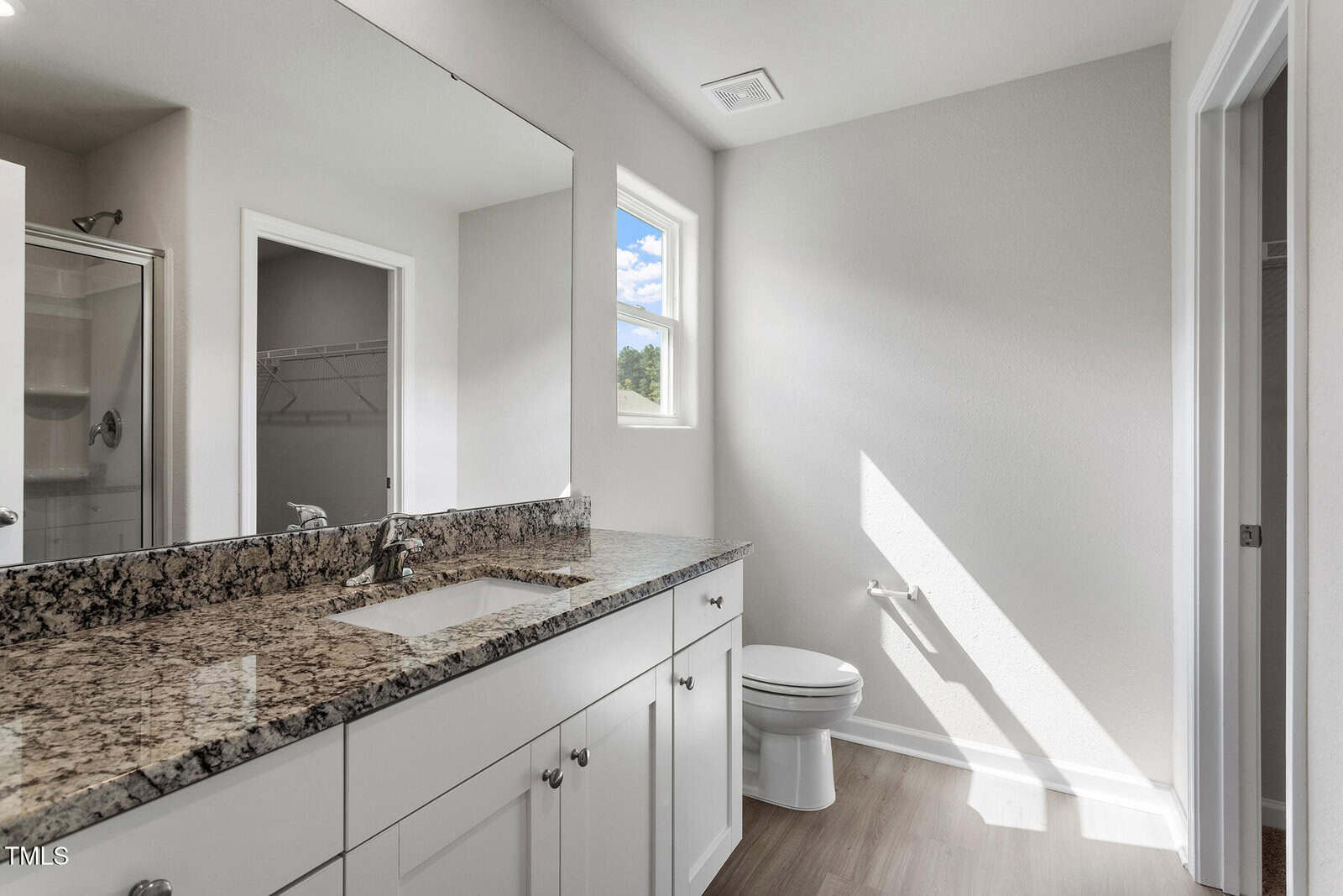 A modern bathroom with a granite countertop, single sink, white cabinets, a toilet, and a window letting in natural light. A shower with glass door and a walk-in closet are visible in the background.
