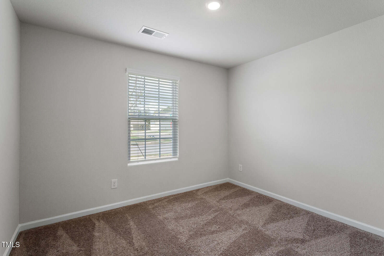 Empty room with beige carpet, white walls, a single window with blinds, and a ceiling light. Sunlight is coming through the window, and there are electrical outlets on the walls.