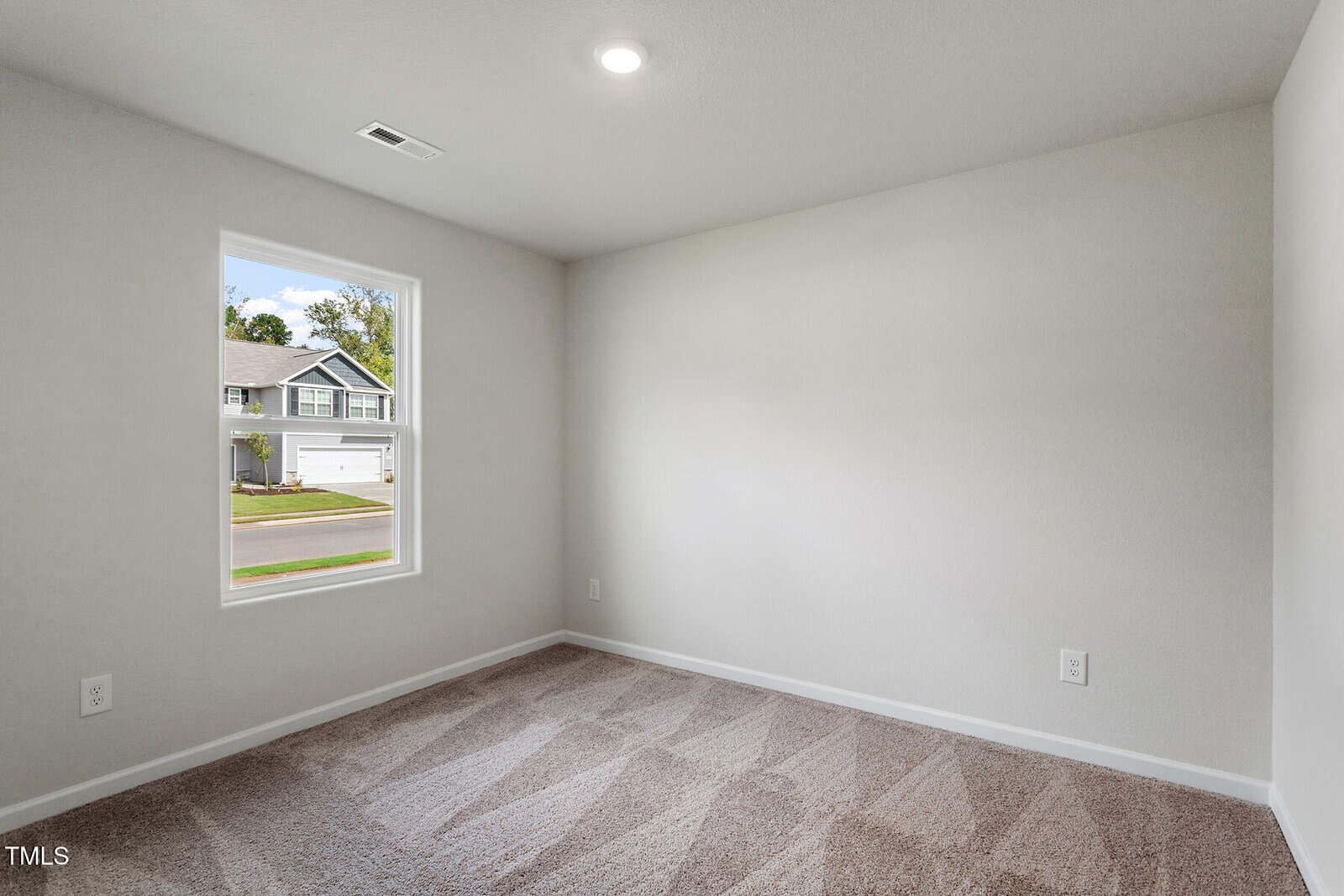 Empty room with light gray walls, beige carpet, a single window showing a suburban street, white baseboards, ceiling light, and electrical outlets visible on two walls.