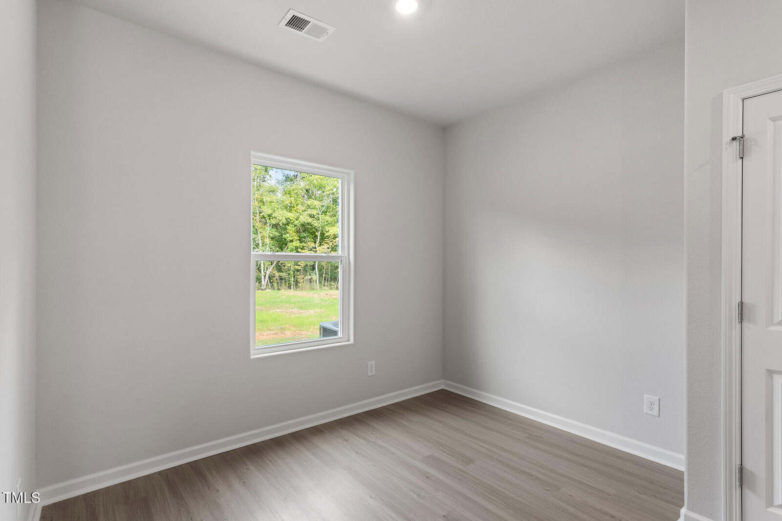 A small, empty room with light gray walls, a single window showing green trees outside, wood-look flooring, and a white door partially visible on the right. Ceiling light and air vent are also visible.