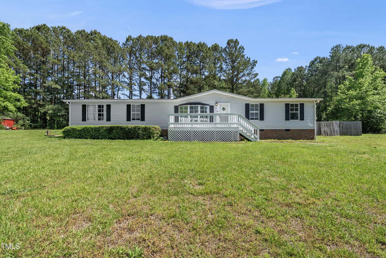 A single-story gray house with white trim and black shutters sits on a grassy lawn, surrounded by tall trees. The house features a small front porch with white railings and steps leading up to the entrance.