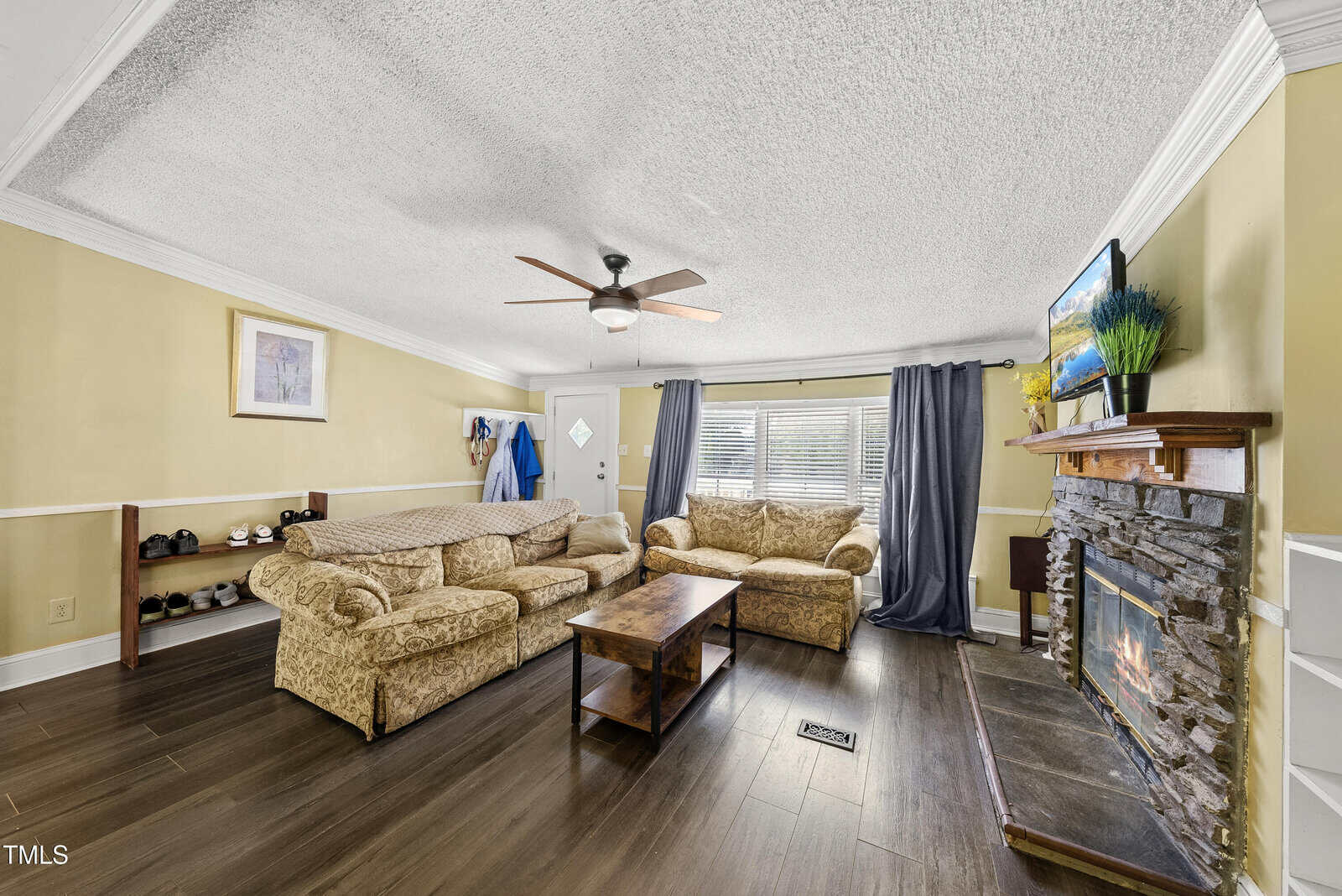A cozy living room with two patterned beige sofas, a wooden coffee table, a stone fireplace with a TV above it, dark wood floors, yellow walls, and a ceiling fan. Coats and shoes are by the door near large curtained windows.