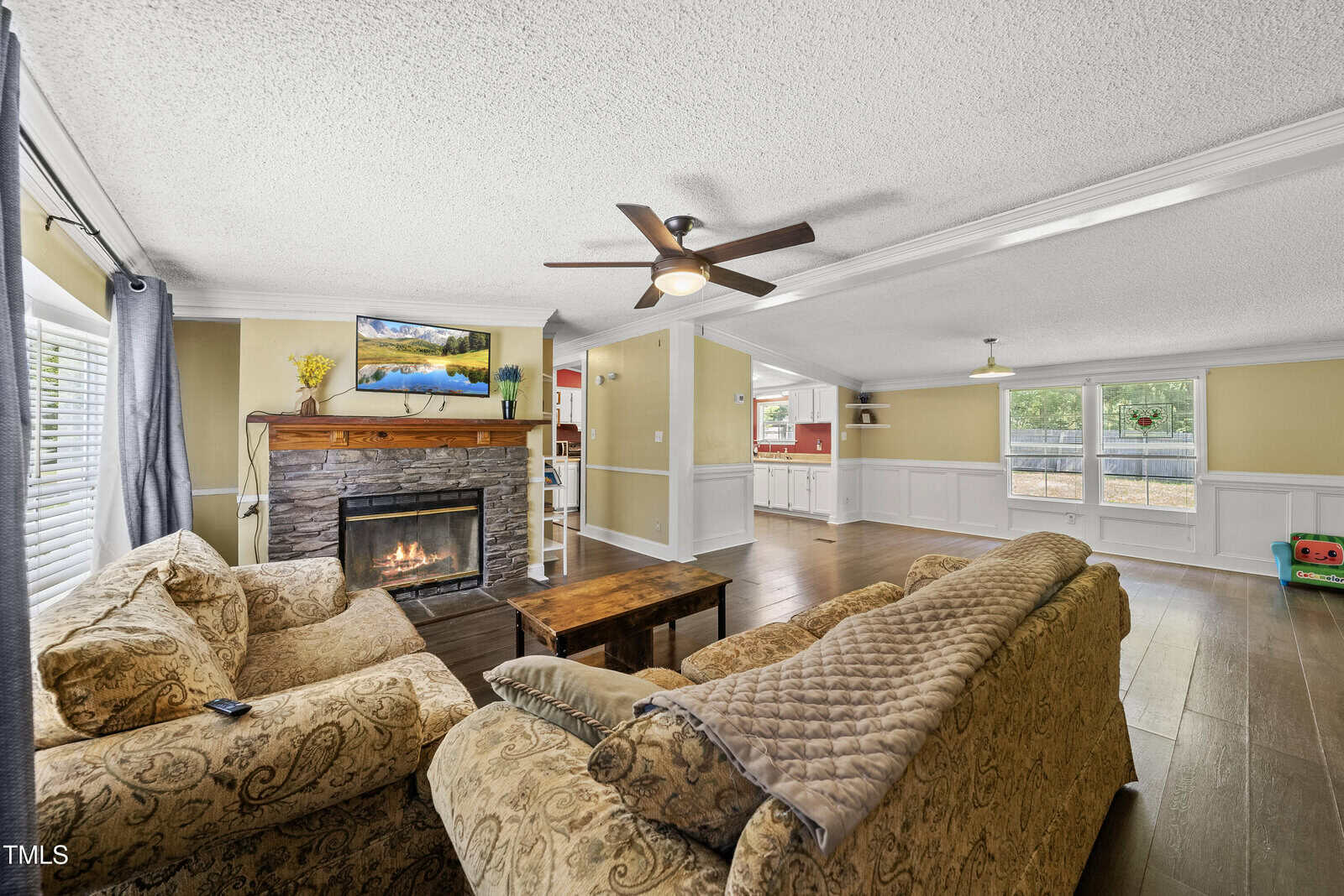 A cozy living room with a stone fireplace, a wall-mounted TV, two patterned sofas, a wooden coffee table, large windows with white trim, and an open view into a bright kitchen and dining area.