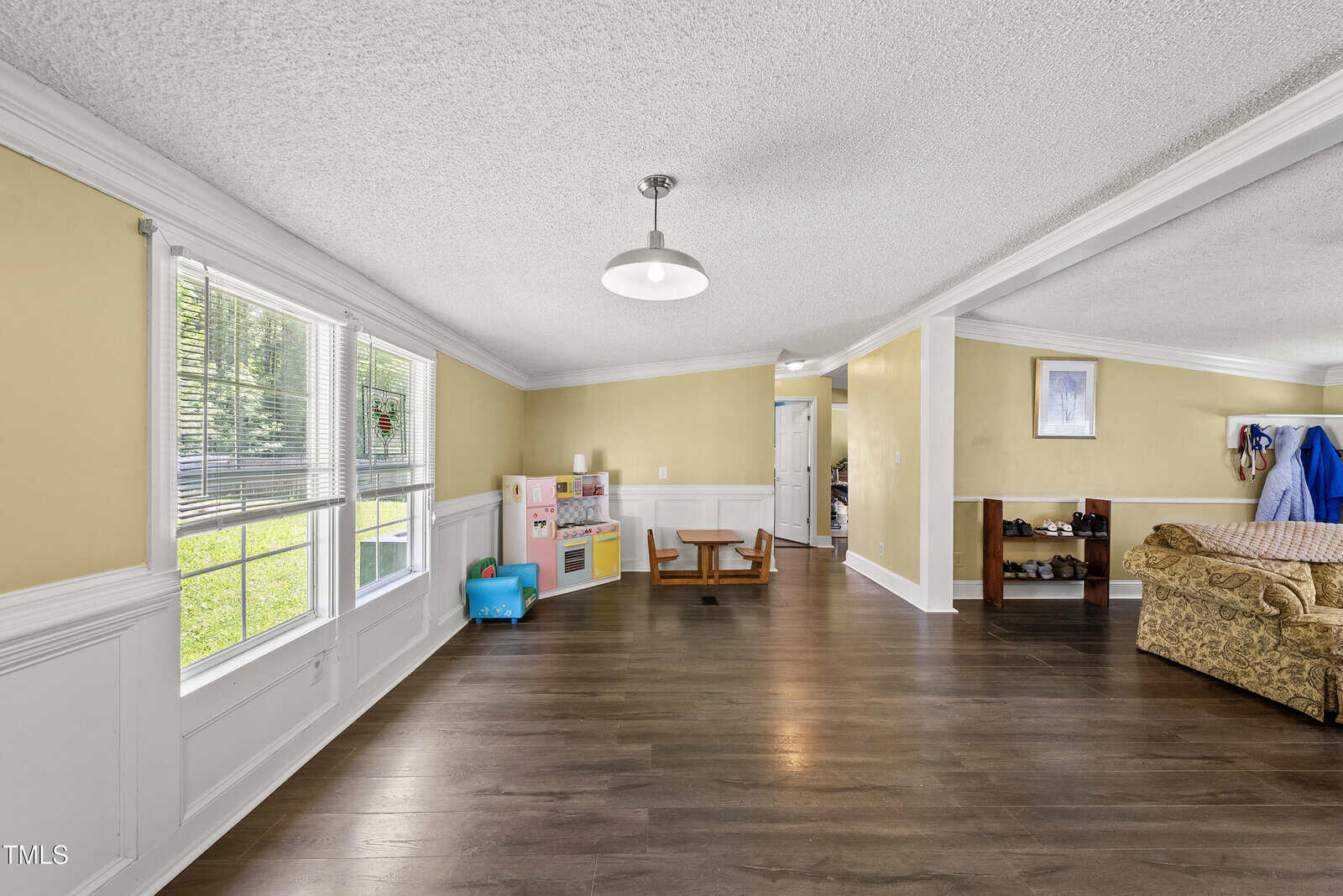 Spacious room with yellow walls, white trim, and wood flooring. Large window on the left lets in sunlight. Children’s play kitchen and toys are in the corner. Sofa and shelf are partially visible on the right.