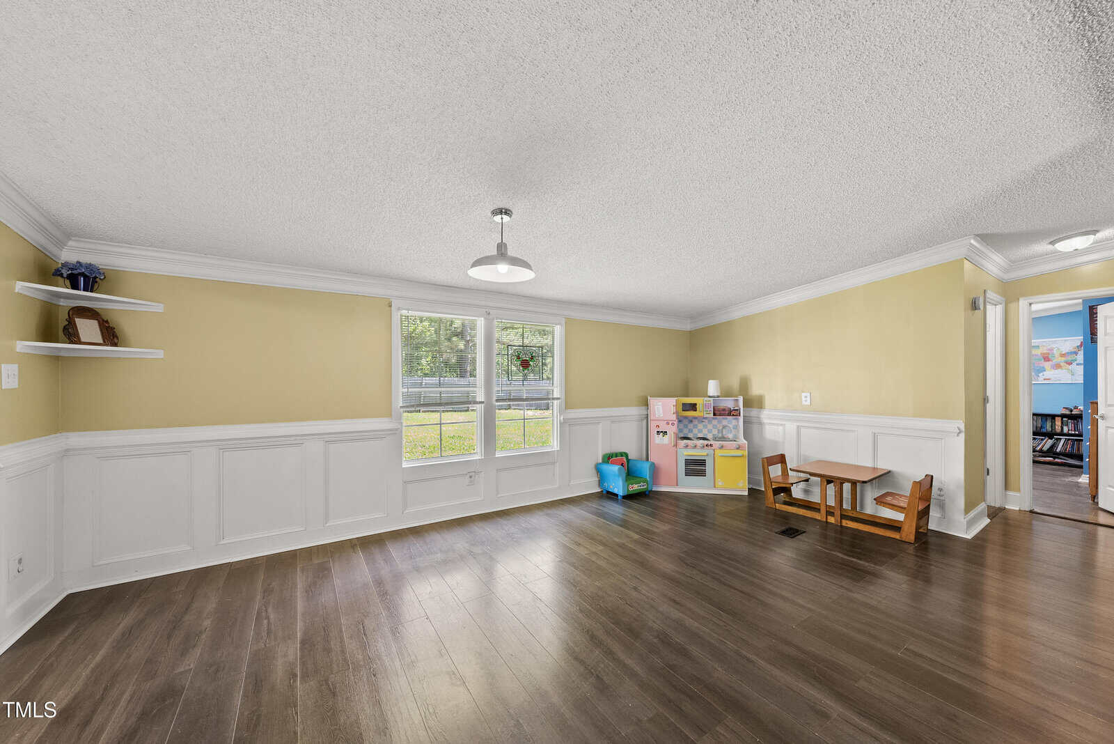 Room with yellow and white walls, dark wood floors, and two windows. There are toy kitchen and table sets on the right side, built-in shelves on the left, and an overhead light fixture in the center.