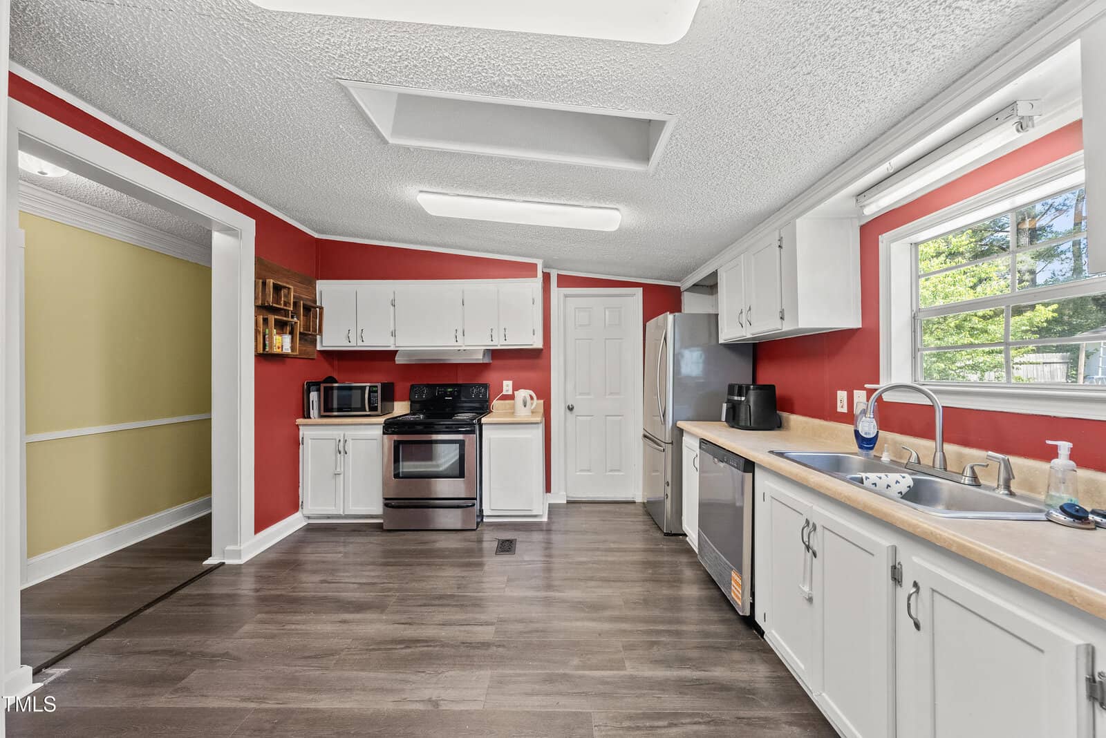 Kitchen with red walls, white cabinets, stainless steel appliances, wood-look flooring, and a window above the sink. Countertops are light-colored, and there are various small kitchen appliances on the counters.