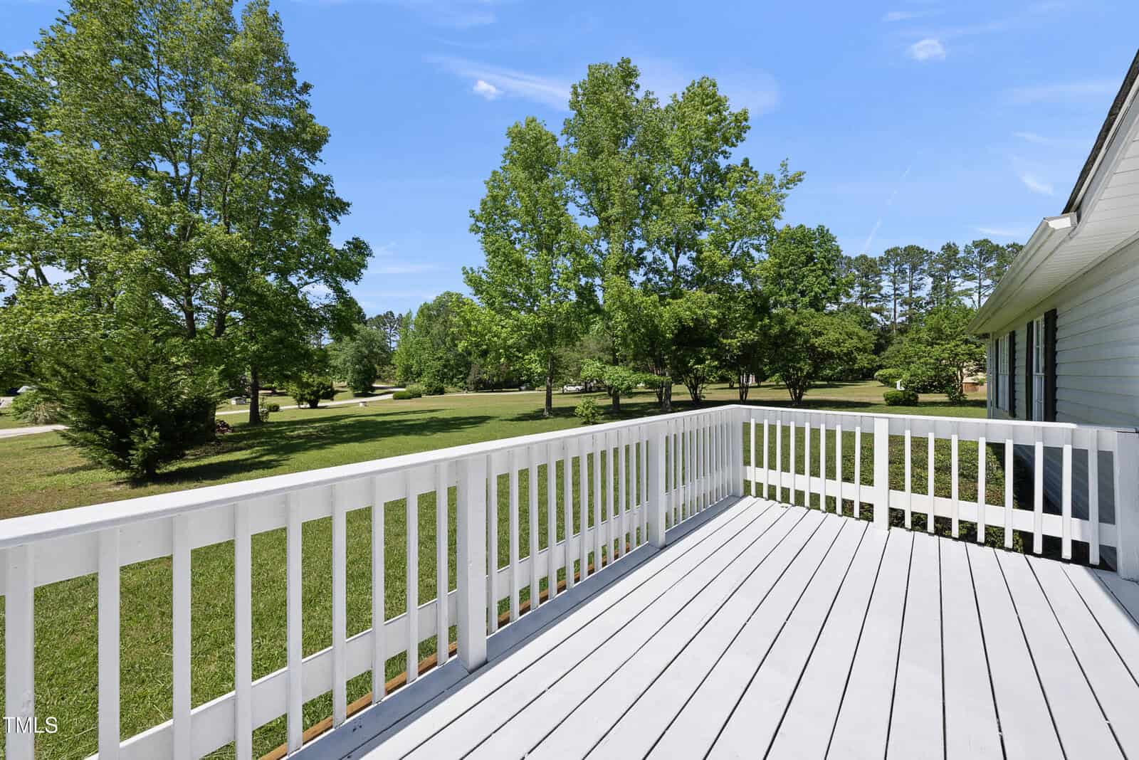 A white wooden deck with railing overlooks a spacious, green lawn with several large trees under a clear blue sky. The side of a light-colored house is visible on the right.