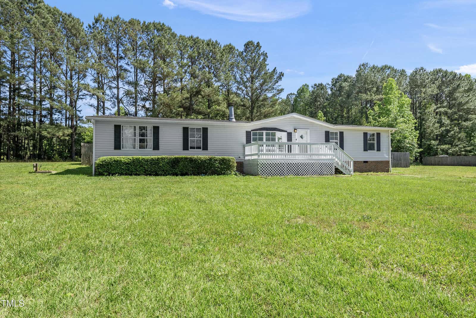 A white, single-story house with black shutters and a raised front porch sits on a large, grassy lawn. Tall trees form a wooded backdrop under a bright blue sky.
