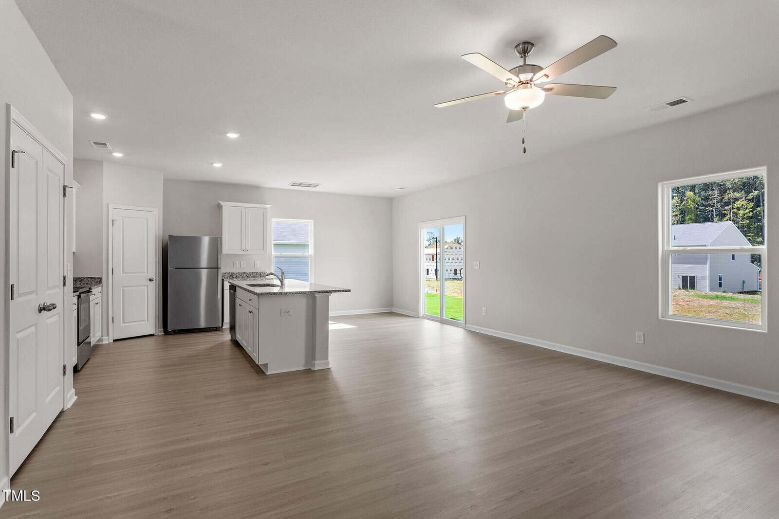 Spacious open-concept kitchen and living area with light wood flooring, white cabinets, stainless steel appliances, ceiling fan, and large windows providing natural light.