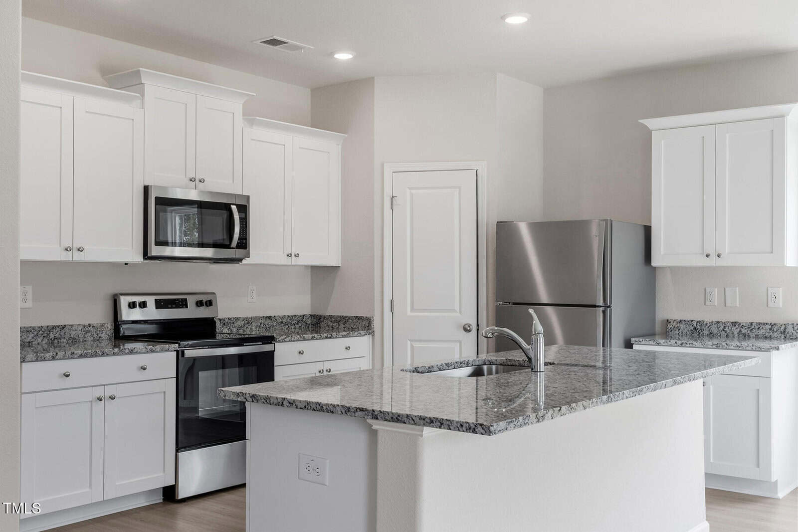 Modern kitchen with white cabinets, granite countertops, stainless steel appliances, an island with a sink and faucet, an electric stove with oven, microwave, and a refrigerator against a light-colored wall.