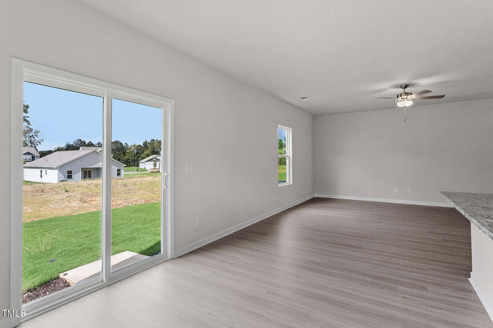 A spacious, empty room with light wood flooring, white walls, a ceiling fan, a window, and a sliding glass door that opens to a grassy backyard and view of neighboring houses.