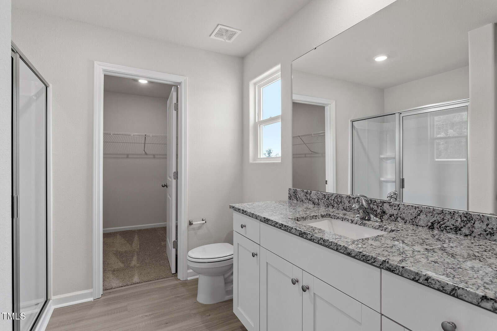 A modern bathroom with light walls, a granite countertop double-sink vanity, a toilet, a shower with glass doors, a window, and a door leading to a walk-in closet with shelving.