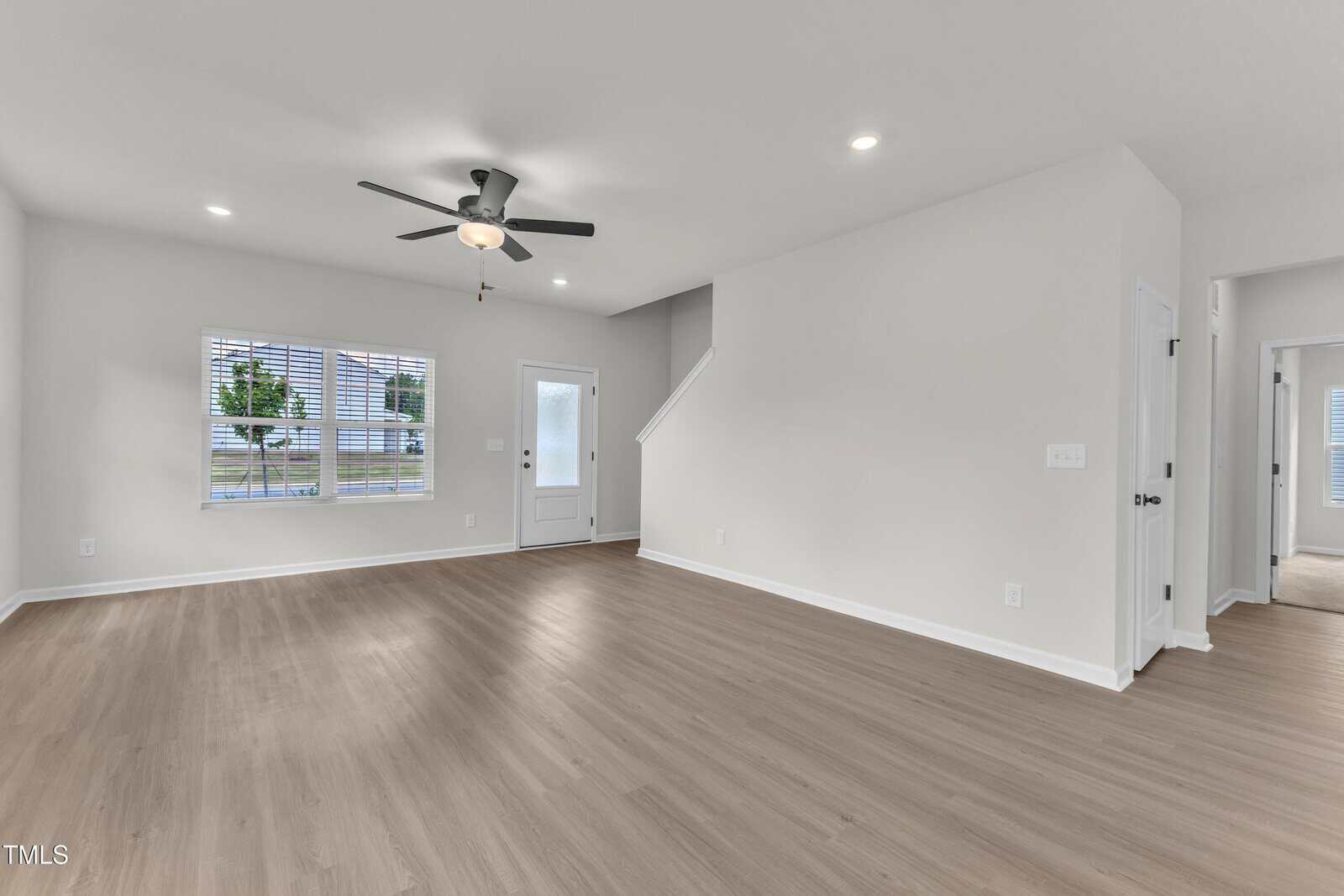 A spacious, empty living room with light wood flooring, white walls, a ceiling fan with light, recessed lighting, a window, a door leading outside, and a staircase to the upper floor.