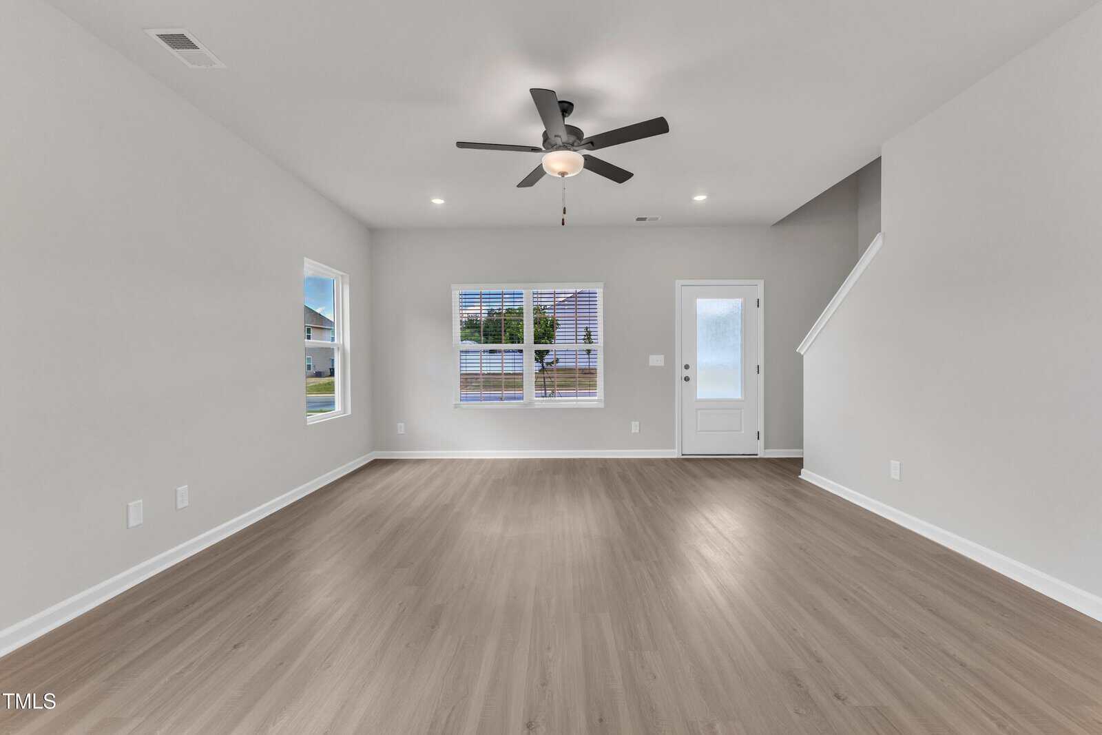 A spacious, empty living room with light wood flooring, white walls, a ceiling fan with lights, two windows, a white door with glass panel, and a staircase leading upward.