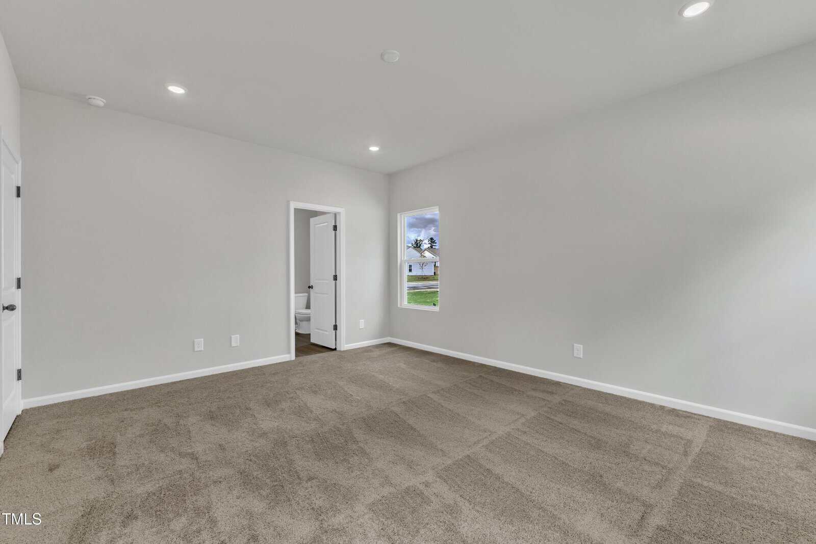 Empty room with beige carpet, white walls, a single window, and a doorway leading to a bathroom. Recessed ceiling lights are on, and there is natural light coming through the window.