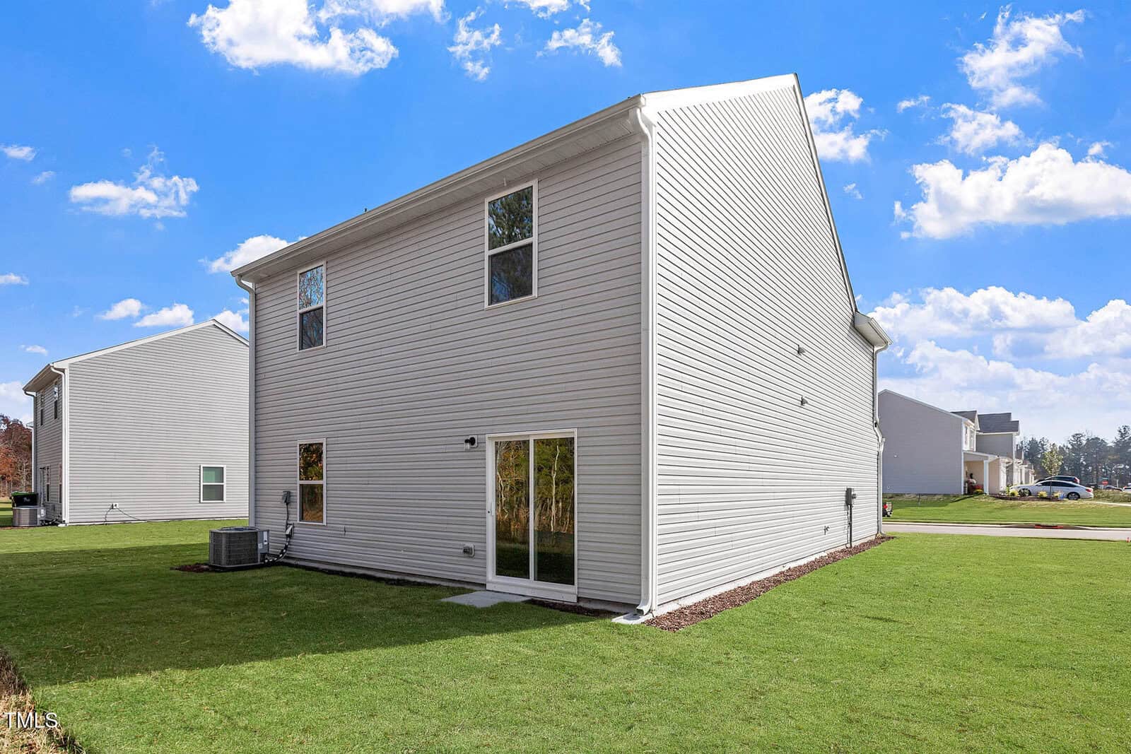 A two-story beige house with vinyl siding and several windows sits on a well-maintained lawn. There is a glass sliding door at the back and an air conditioning unit on the side. The sky is clear with scattered clouds.