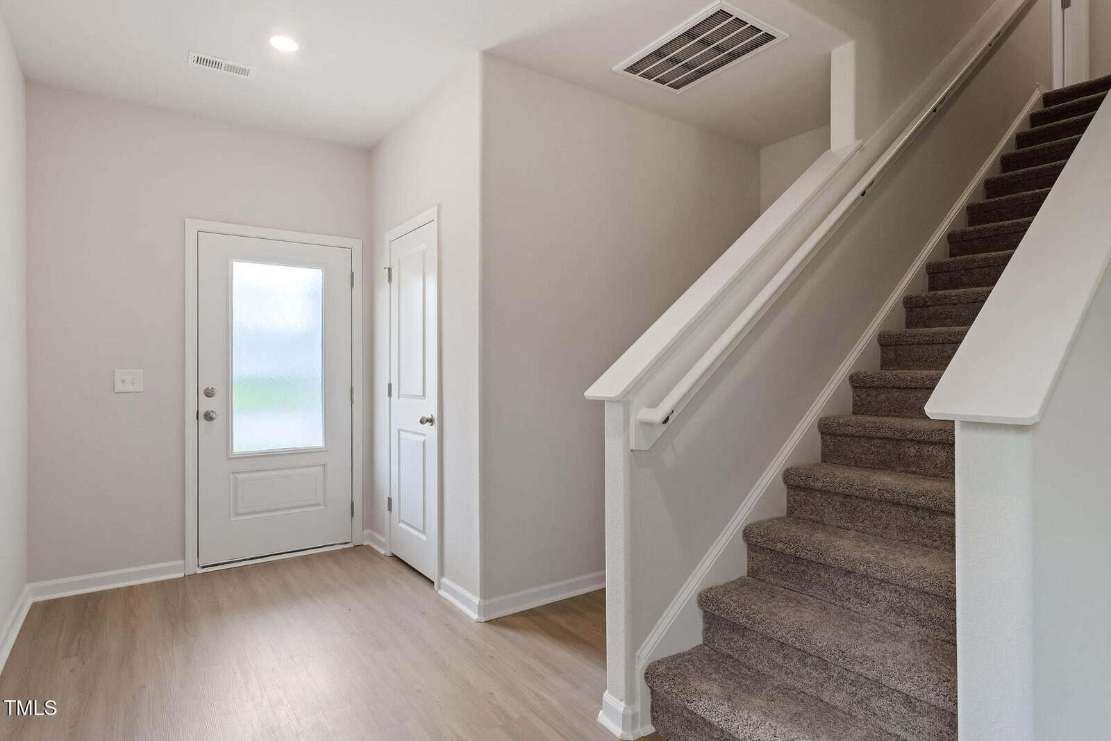 A modern entryway with a white front door, a closet door, light wood flooring, and a carpeted staircase with a white railing leading to the second floor. Walls are painted light beige.