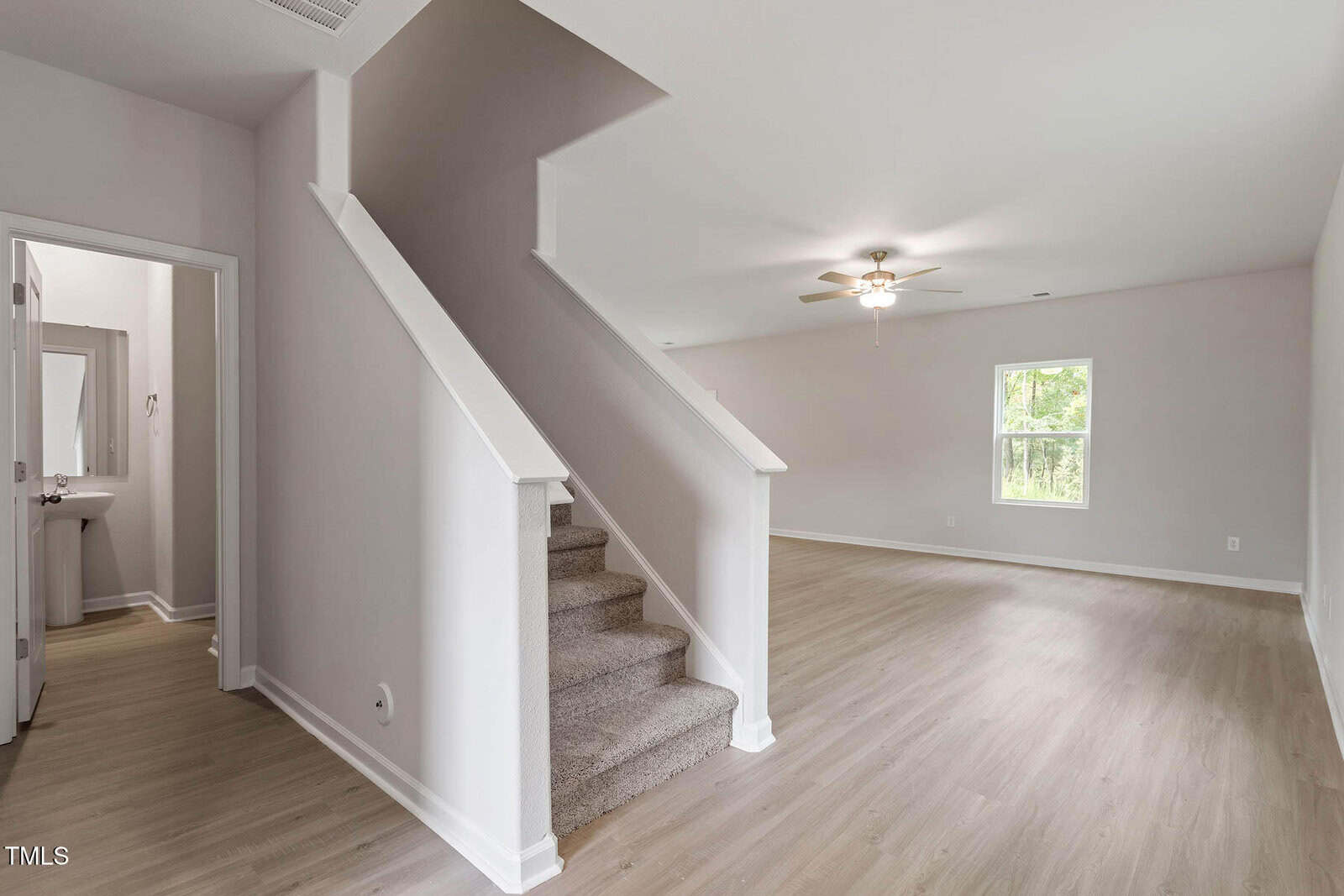 A modern, empty living room with light wood flooring, white walls, a window, a ceiling fan with light, and a staircase with carpeted steps leading upward. A door to a bathroom is visible on the left.