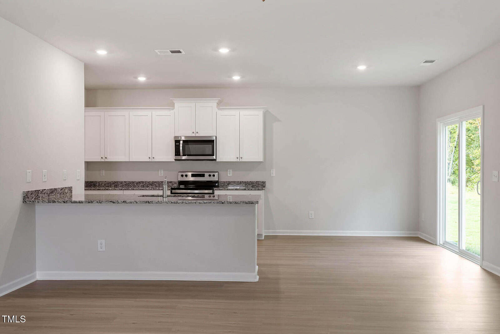 A modern kitchen with white cabinets, stainless steel appliances, granite countertops, and a breakfast bar opens to a spacious, empty living area with wood flooring and a glass patio door leading outside.