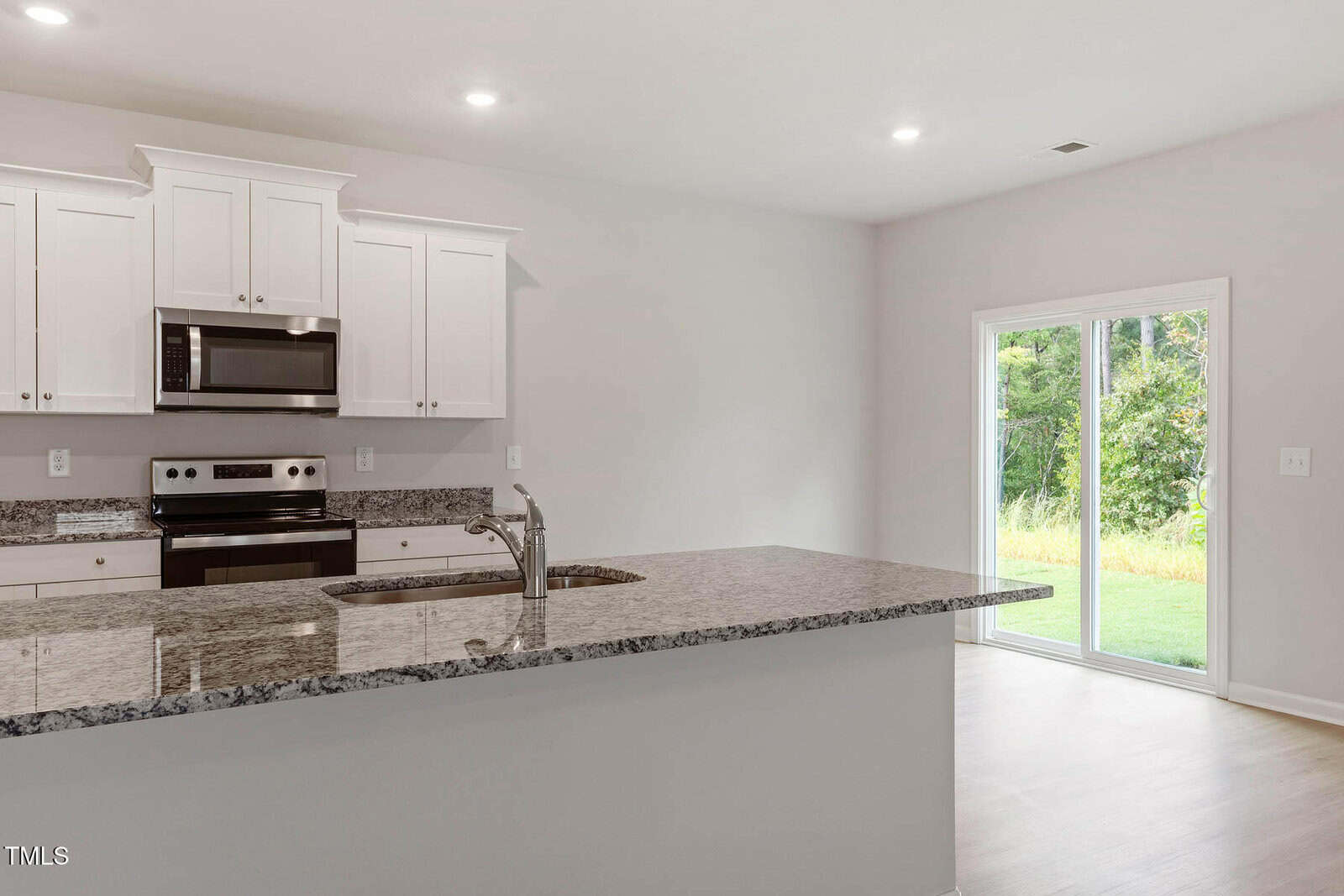 A modern kitchen with white cabinets, stainless steel appliances, a granite countertop island with a sink, and sliding glass doors leading to a grassy backyard. Bright natural light fills the space.