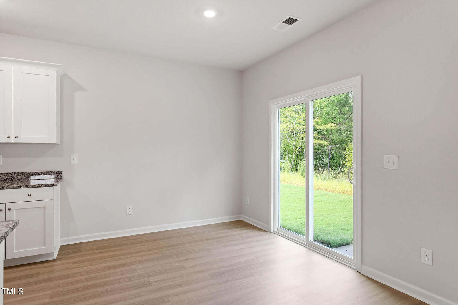 A bright, unfurnished room with light wood flooring, white walls, a sliding glass door leading to a green yard, and part of a kitchen counter with white cabinets and granite countertop visible on the left.