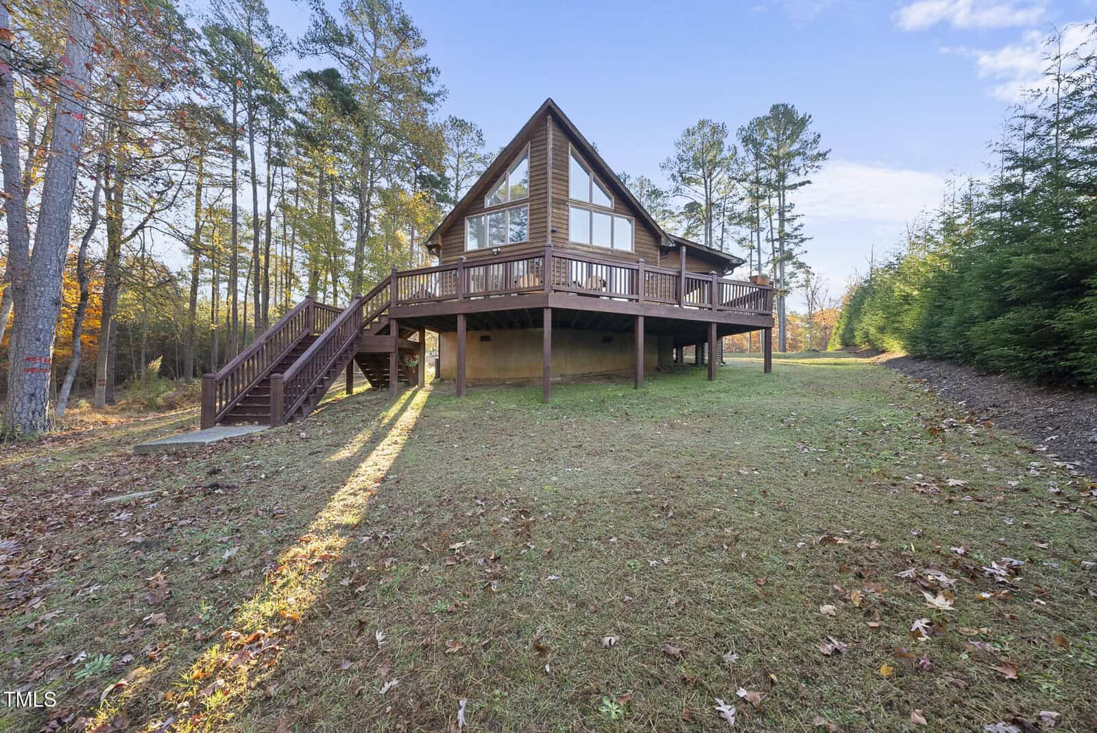 A wooden A-frame cabin with a large wraparound deck stands elevated on stilts among tall trees, surrounded by a grassy lawn with scattered autumn leaves under a clear blue sky.