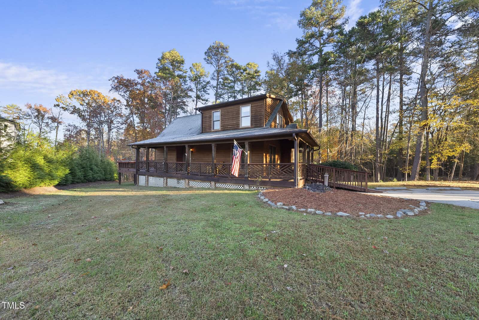 A brown wooden house with a wraparound porch and American flag, surrounded by trees and a large grassy yard, under a clear blue sky.