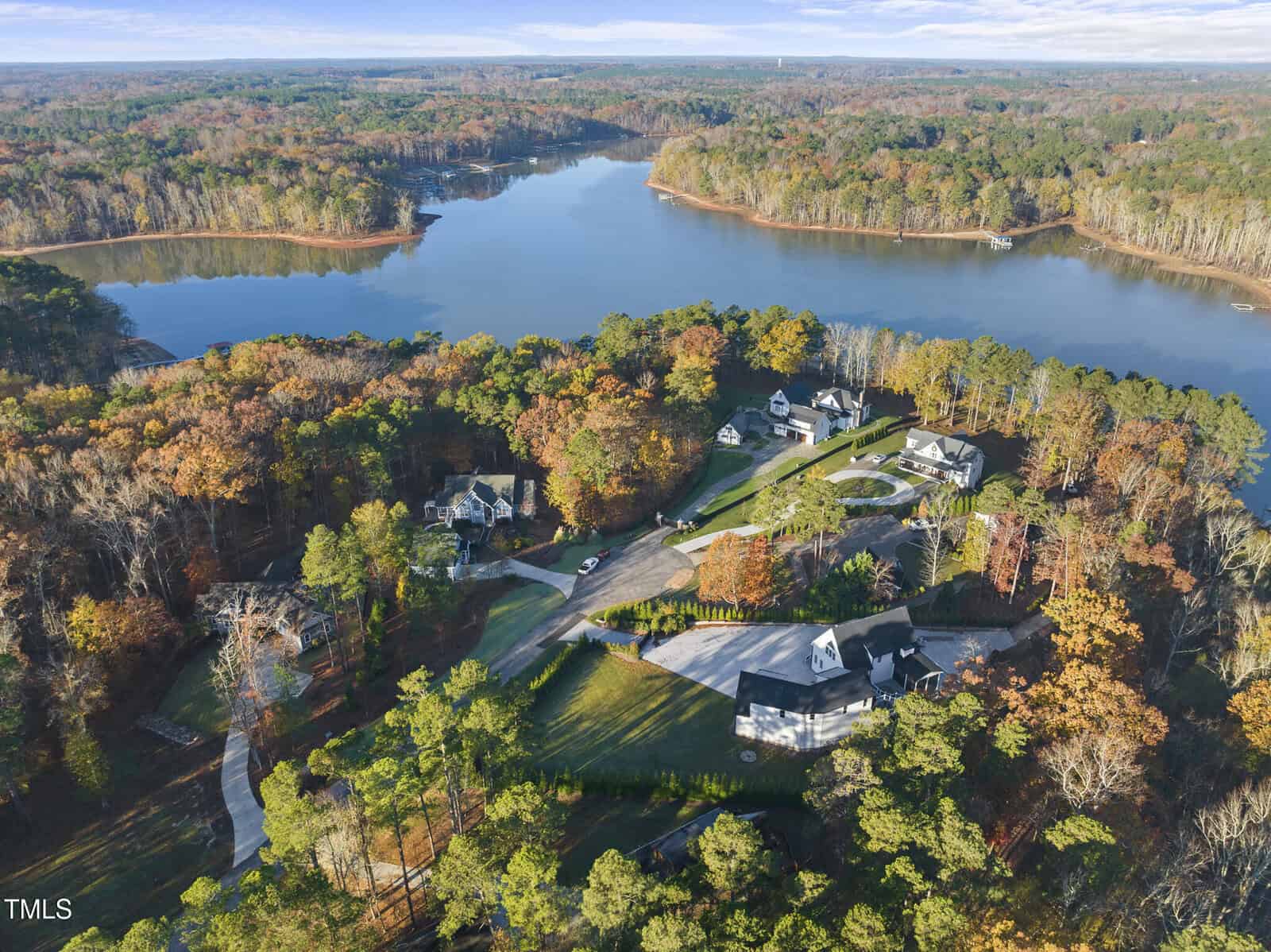 Aerial view of a lakeside neighborhood with several large houses surrounded by autumn trees, set beside a winding lake with wooded shores under a partly cloudy sky.