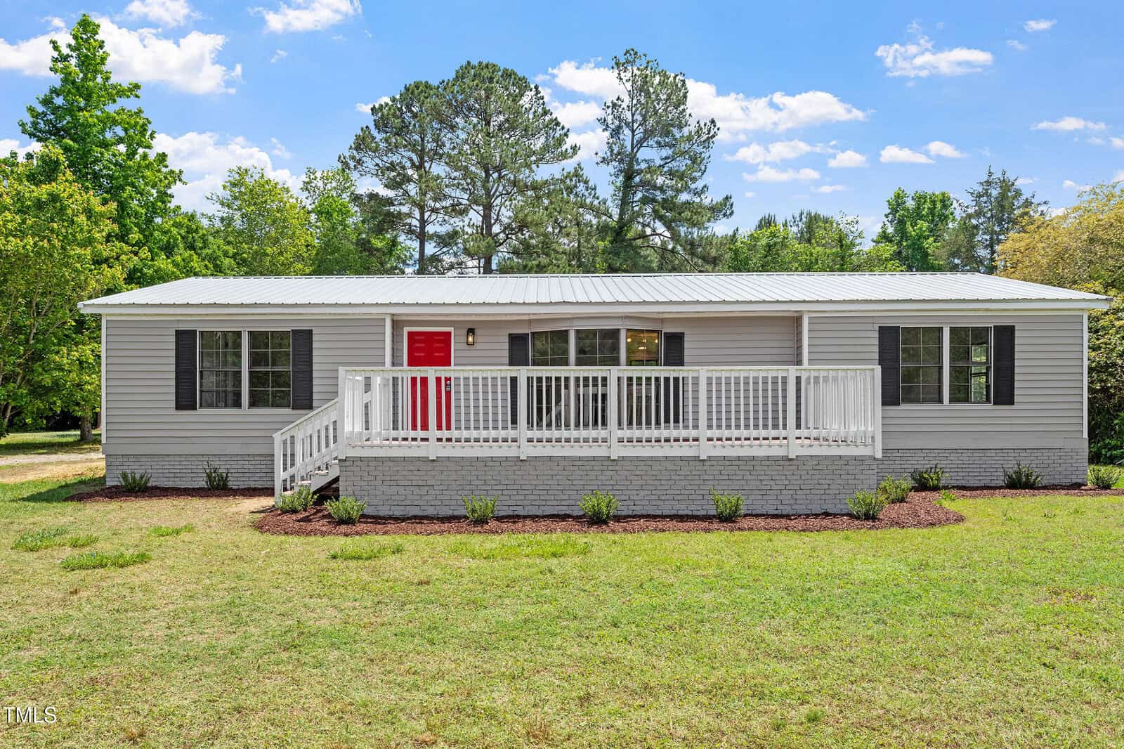 Single-story gray house with black shutters, a bright red front door, and a white-railed porch. The yard is grassy with a few small shrubs, and there are tall trees in the background under a blue sky with clouds.